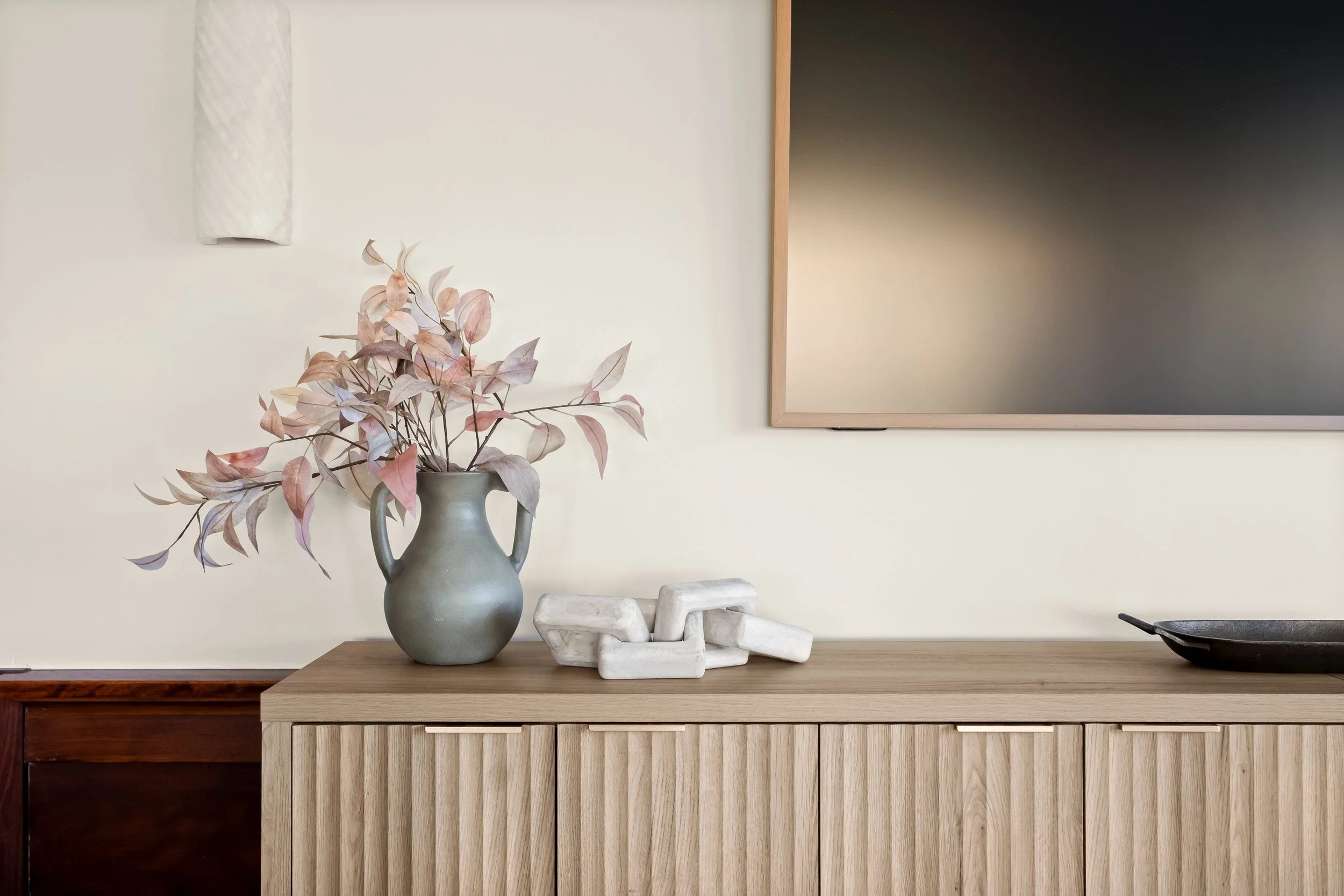 A wooden sideboard with a light wood finish, topped with a gray vase containing pinkish leaves, white decorative stones, and a black shallow bowl. There is a large flat black frame or mirror on the light-colored wall above the sideboard.