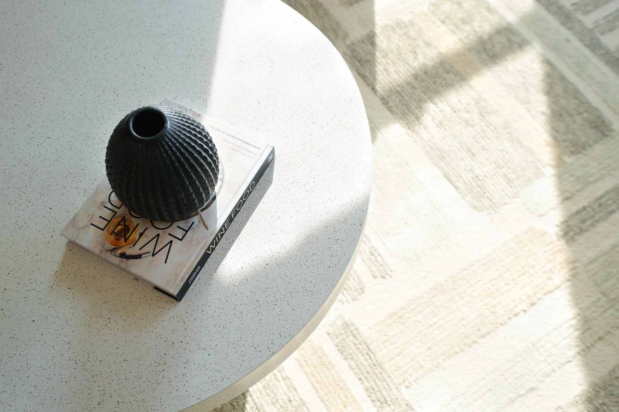 Black textured vase placed on top of a magazine on a white tabletop with a natural fiber rug and patterned rug on the floor nearby.