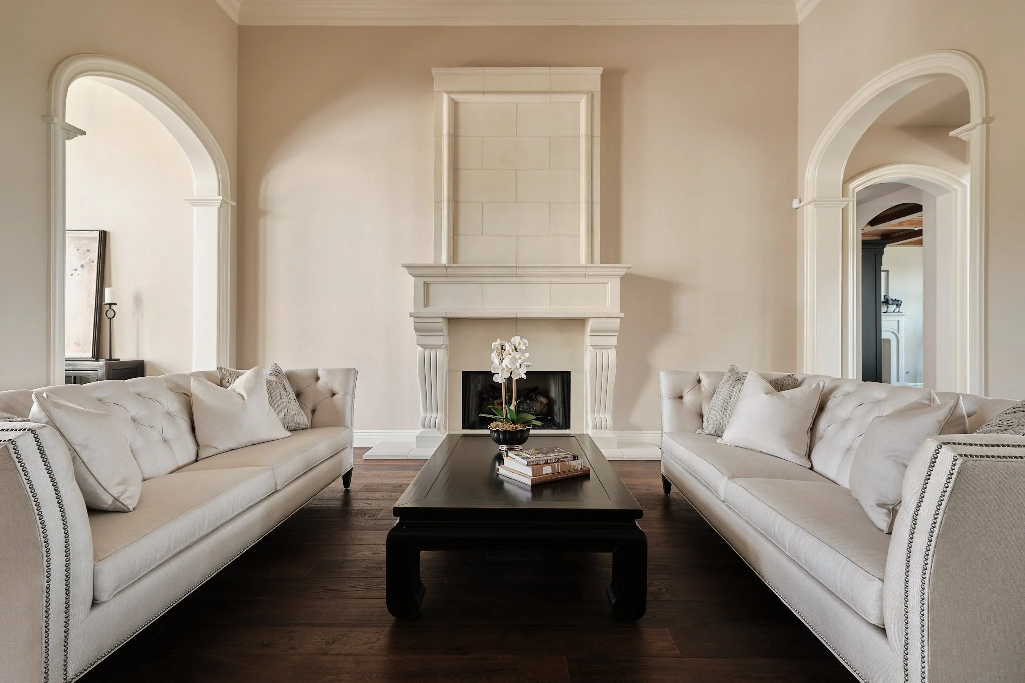 Living room with beige walls, white fireplace, two white upholstered sofas facing each other, dark wood coffee table with books, and arched doorways on each side.