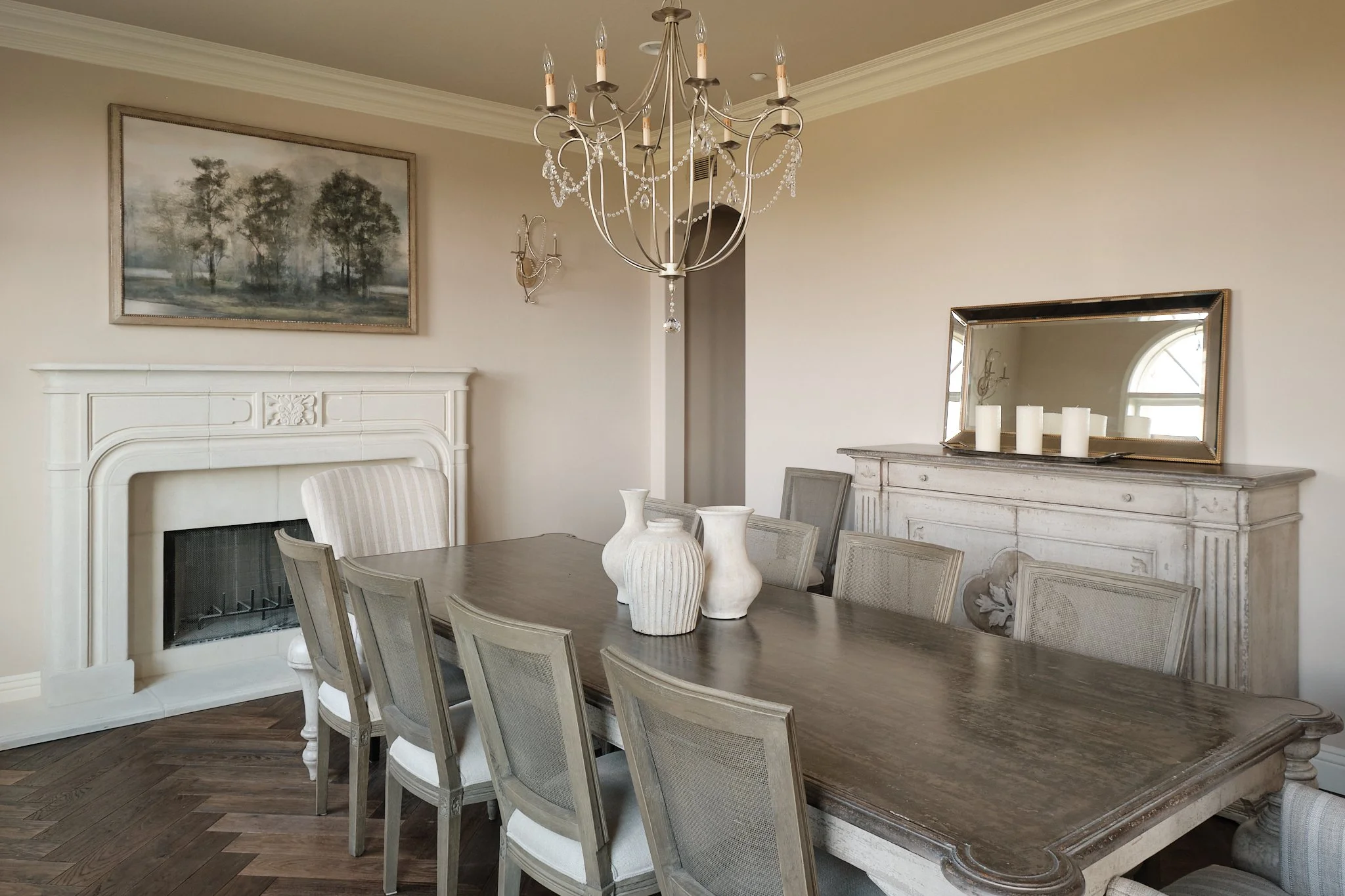 A dining room with a rectangular wooden table, seven chairs, a white fireplace mantel, a chandelier, a large mirror, a painting of trees, and decorative vases on a vintage white sideboard.