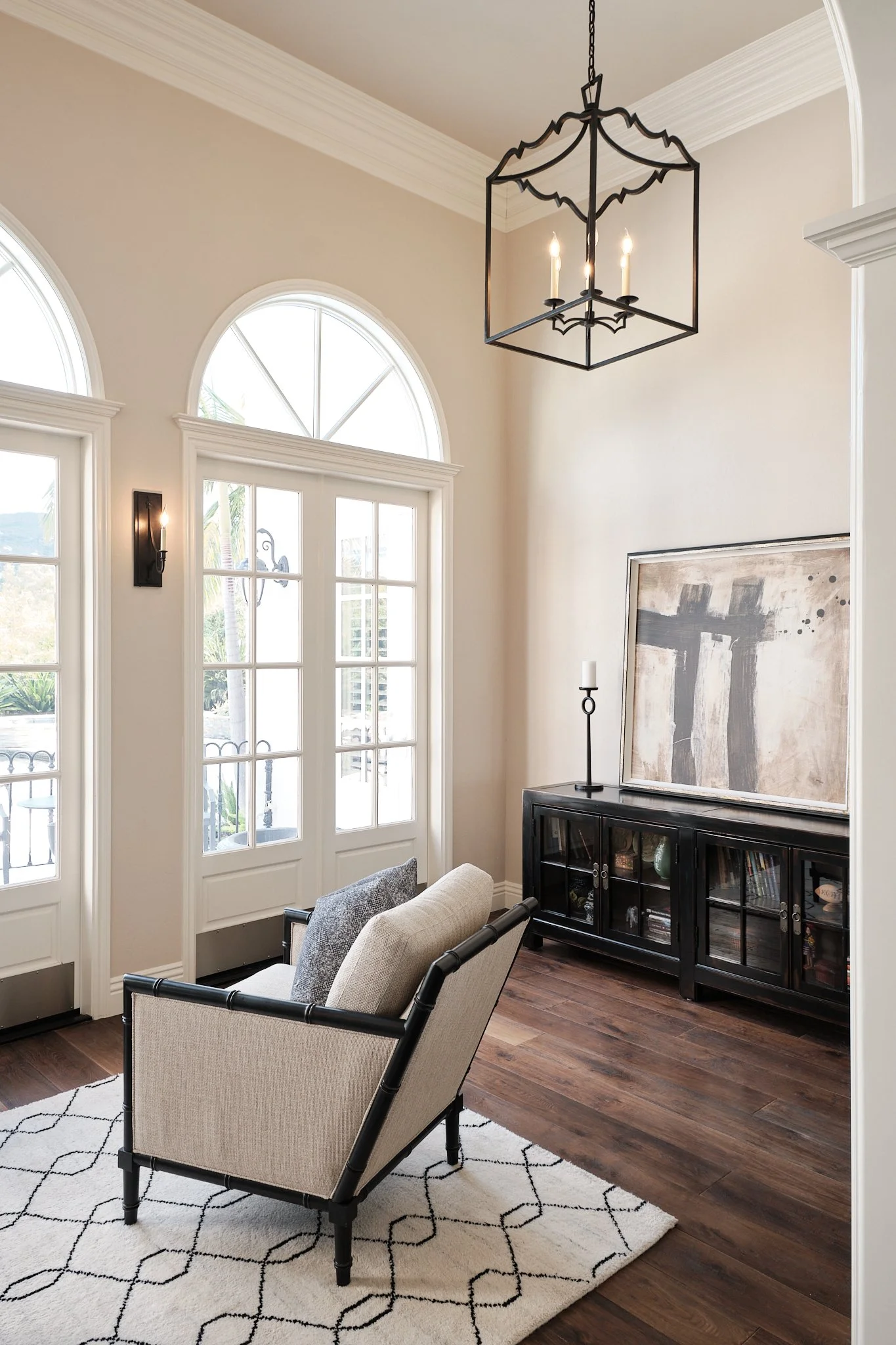 A living room with white walls, large windows, a black wooden cabinet with glass doors, abstract art, a beige chair, a black metal and glass coffee table, a patterned white and black rug, and a black metal chandelier with three candle-style lights.