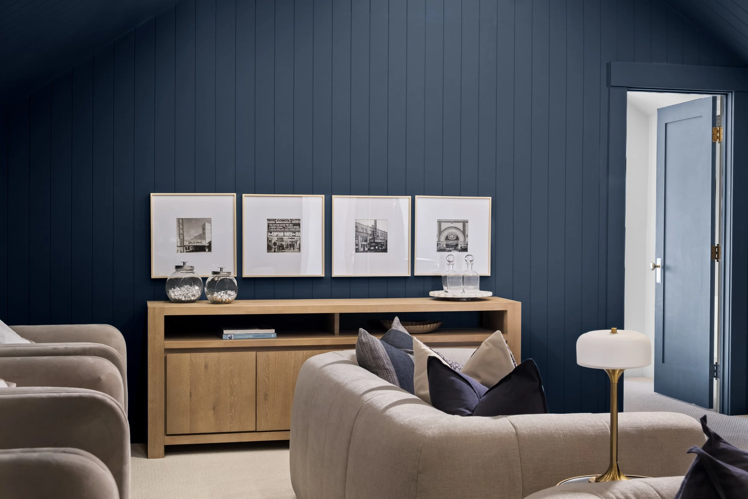 Living room with a navy blue wall, framed black-and-white photographs, a wooden console table with decorative glass jars, beige sofas with pillows, and a white and gold floor lamp near an open door.