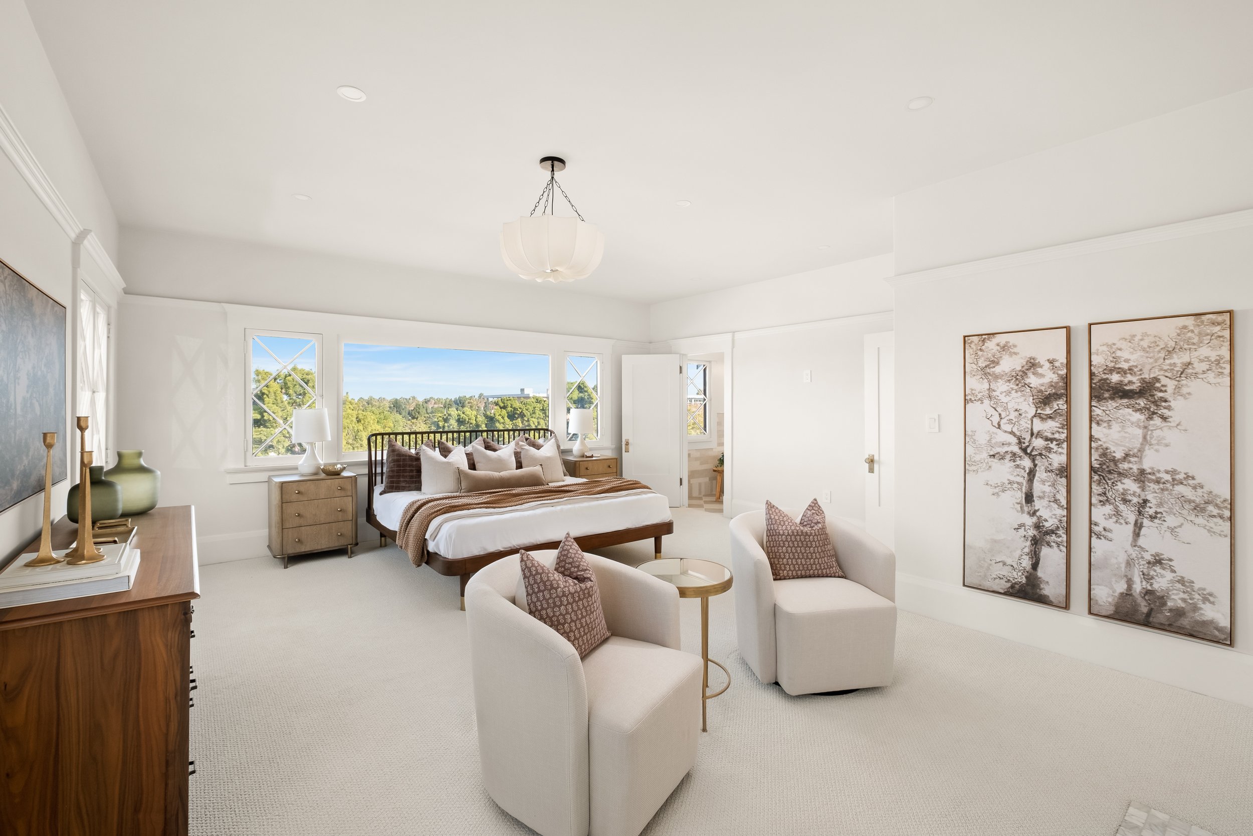 Bright bedroom with large windows overlooking a green landscape, featuring a wooden bed with white and brown bedding, side tables with lamps, and decorative artwork on the wall.