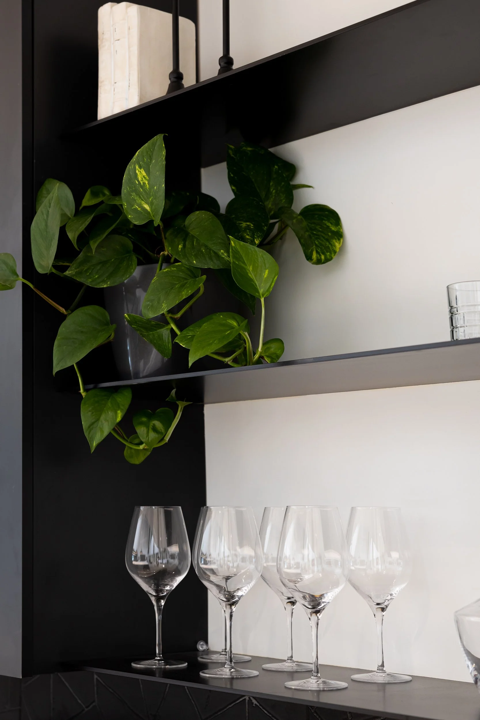 A black shelf with a plant and books on the upper level, glassware including wine glasses on the middle level, and a white wall background.