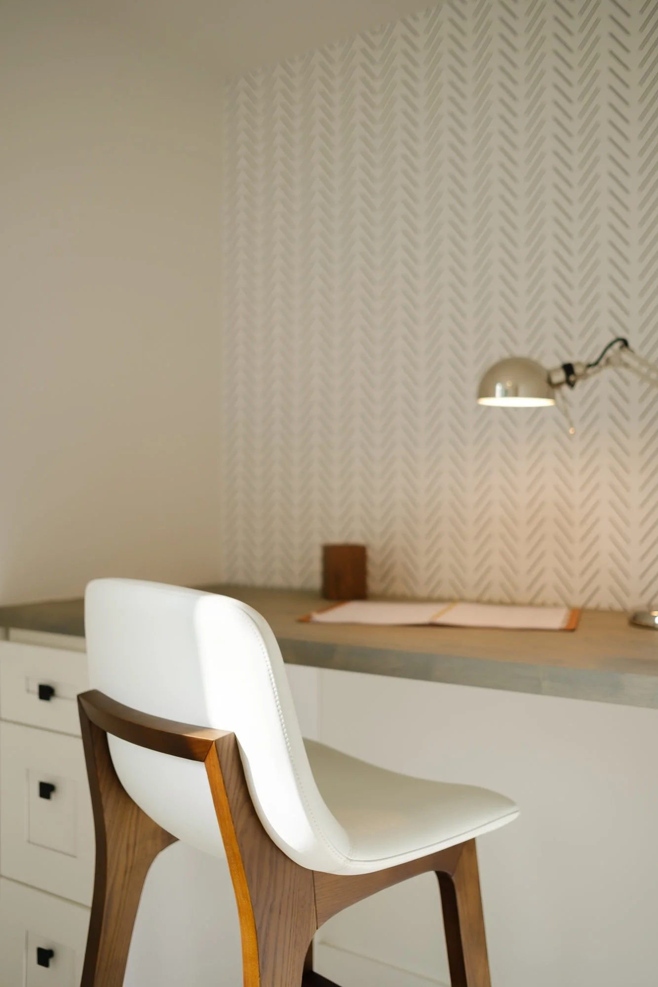 A modern dining or workspace area with a white chair with wooden arms and legs in the foreground. In the background, a desk with a drawer and a small dark handle is visible. On the wall behind, there is textured wallpaper with a geometric pattern, an