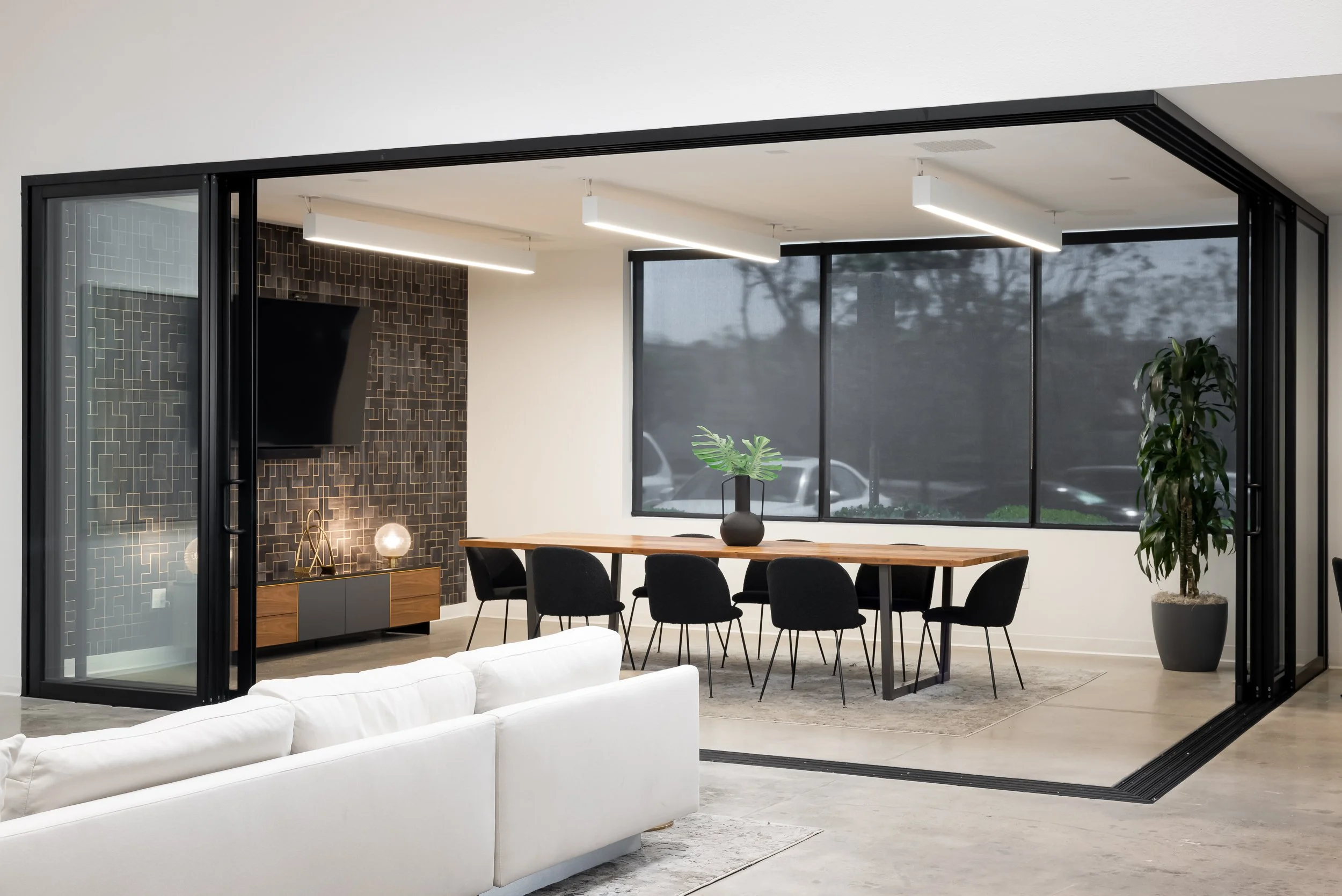 Modern open-concept living and dining area with large windows, a white sofa, a wooden dining table, black chairs, and decorative plants.