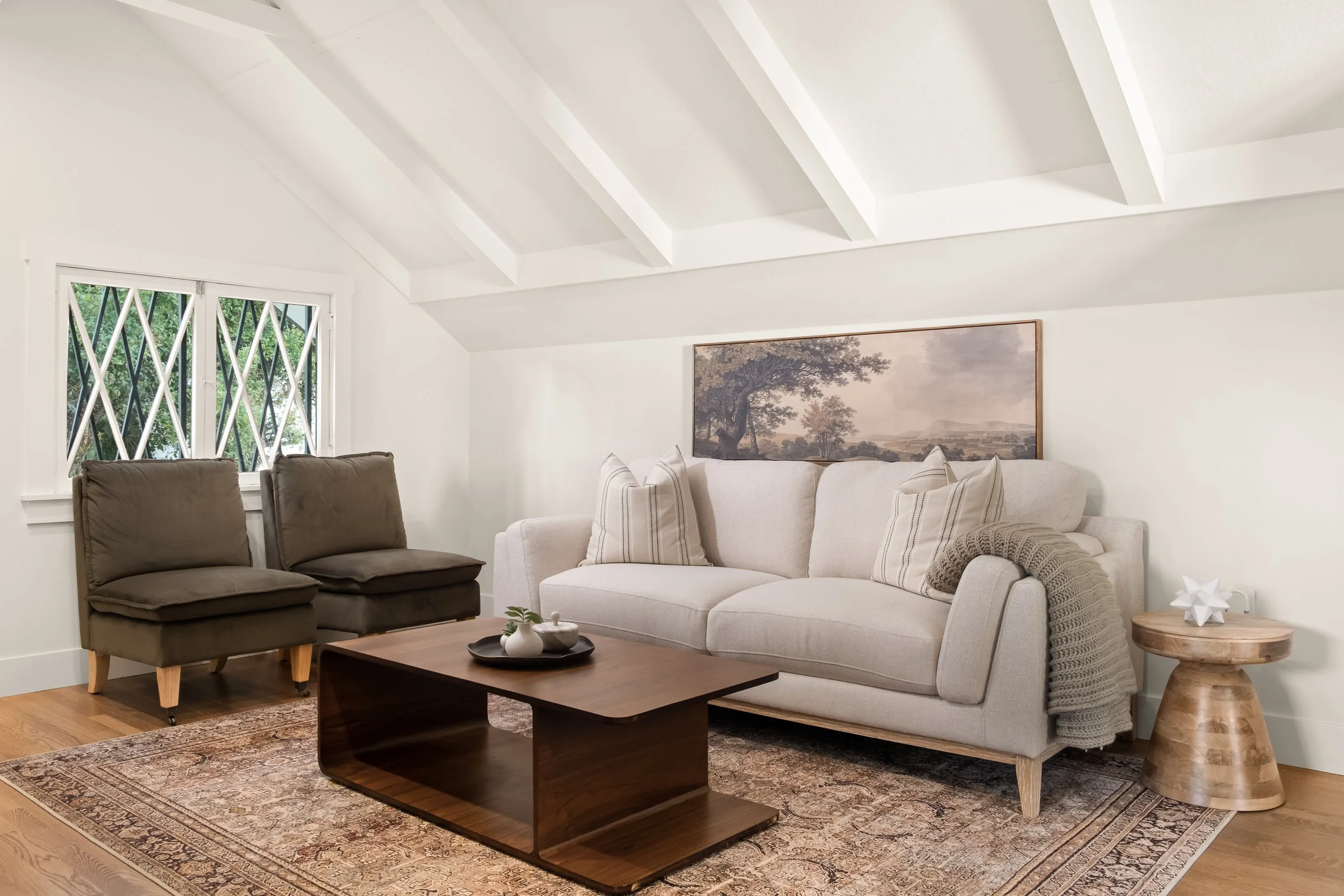 A living room with white walls, a white sofa with pillows, a wooden side table, two brown chairs, a wooden coffee table, a patterned rug, a large landscape painting on the wall, a window with angled panes, and decorative items on the coffee table and