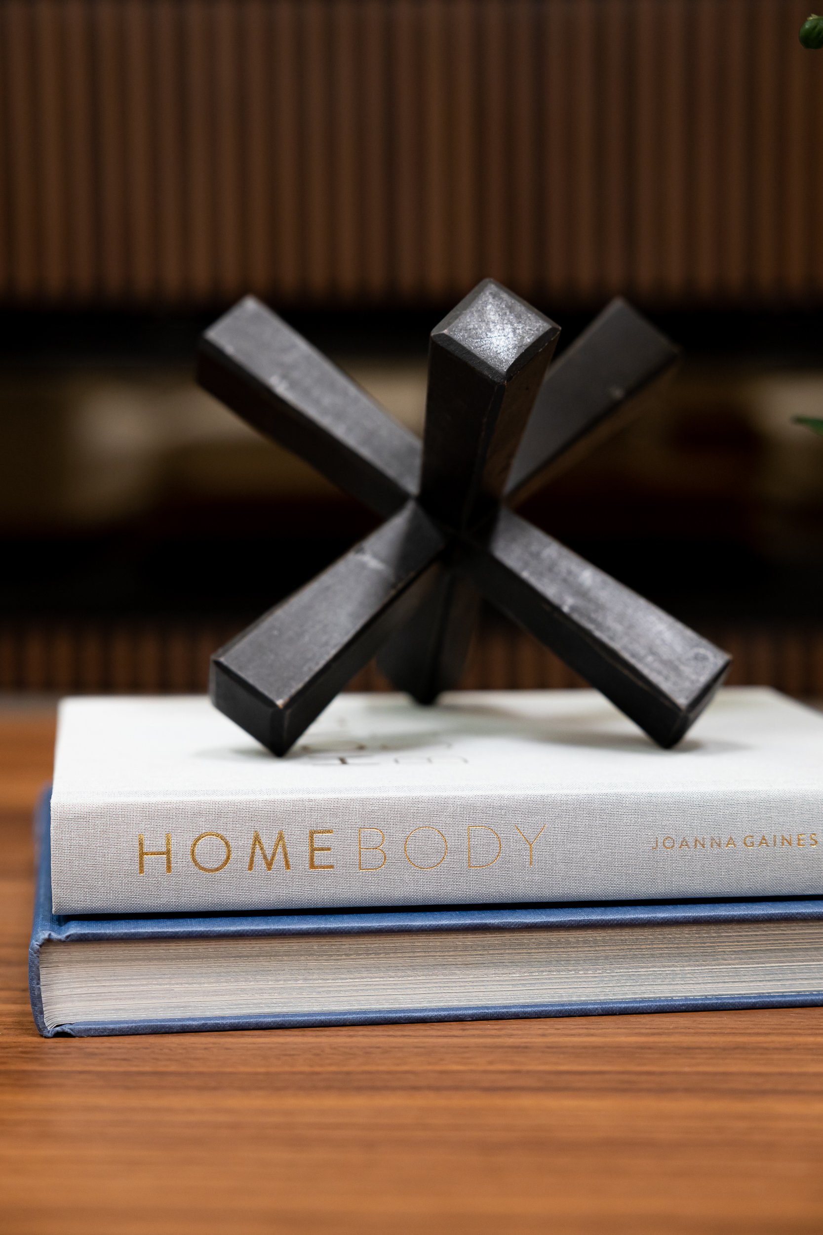 A stack of two books with a black metal sculpture on top, featuring four intersecting rectangular bars in a star shape. The top book has a white cover with the words 'HOME BODY' and the author's name 'JOANNA GAINES' written in gold. The background is