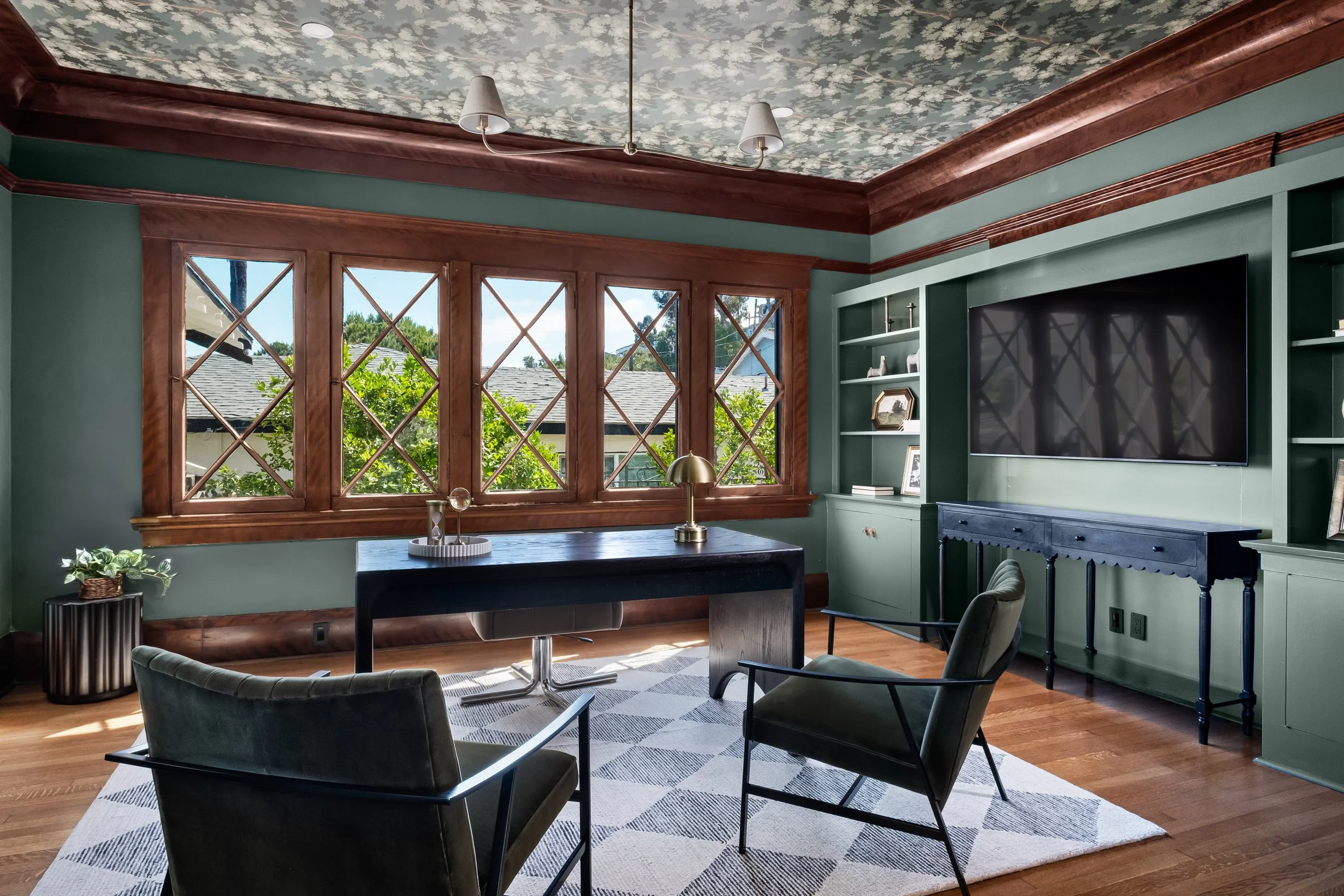 A cozy living room with large wooden-framed windows, green walls, a navy blue cabinet, a wall-mounted flat-screen TV, a small side table, and a patterned area rug. The ceiling has floral wallpaper and two pendant lights.