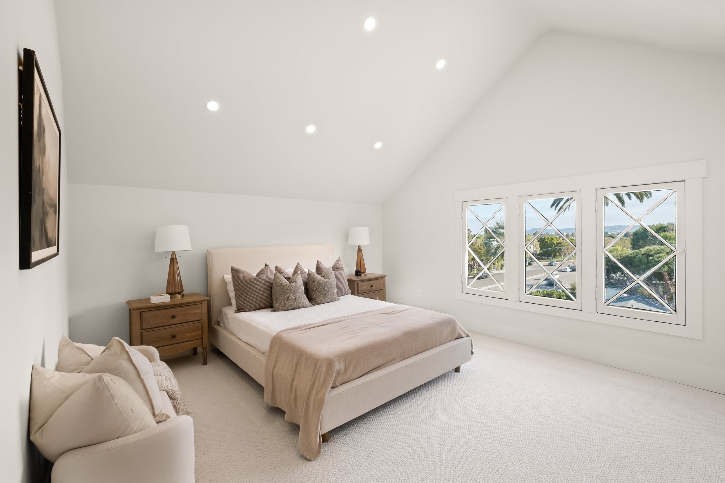 A bright bedroom with white walls and large windows overlooking trees and a street. There is a white bed with beige and patterned pillows, wooden nightstands with white lamps, and a black-framed wall art.