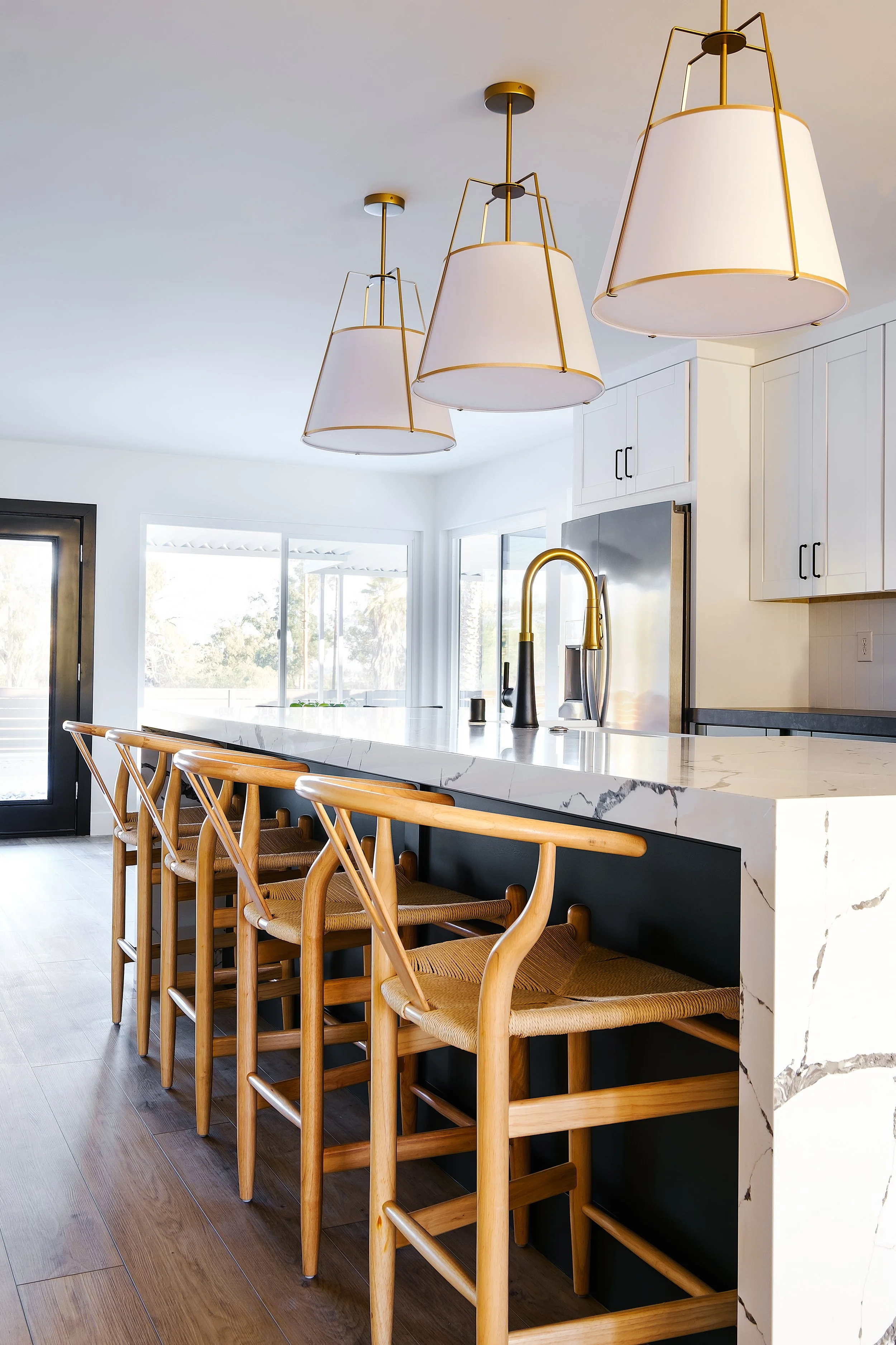 Modern kitchen with white cabinets, marble island, gold pendant lights, wooden bar stools, and large windows.