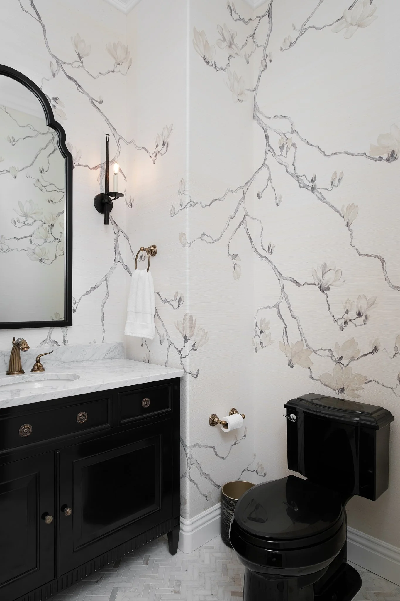 A bathroom with a dark vanity, white marble countertop, and a black modern toilet. The wall has a large white floral wallpaper and a round mirror framed in black. There's a wall-mounted black sconce and a brass towel holder with a white towel. The fl
