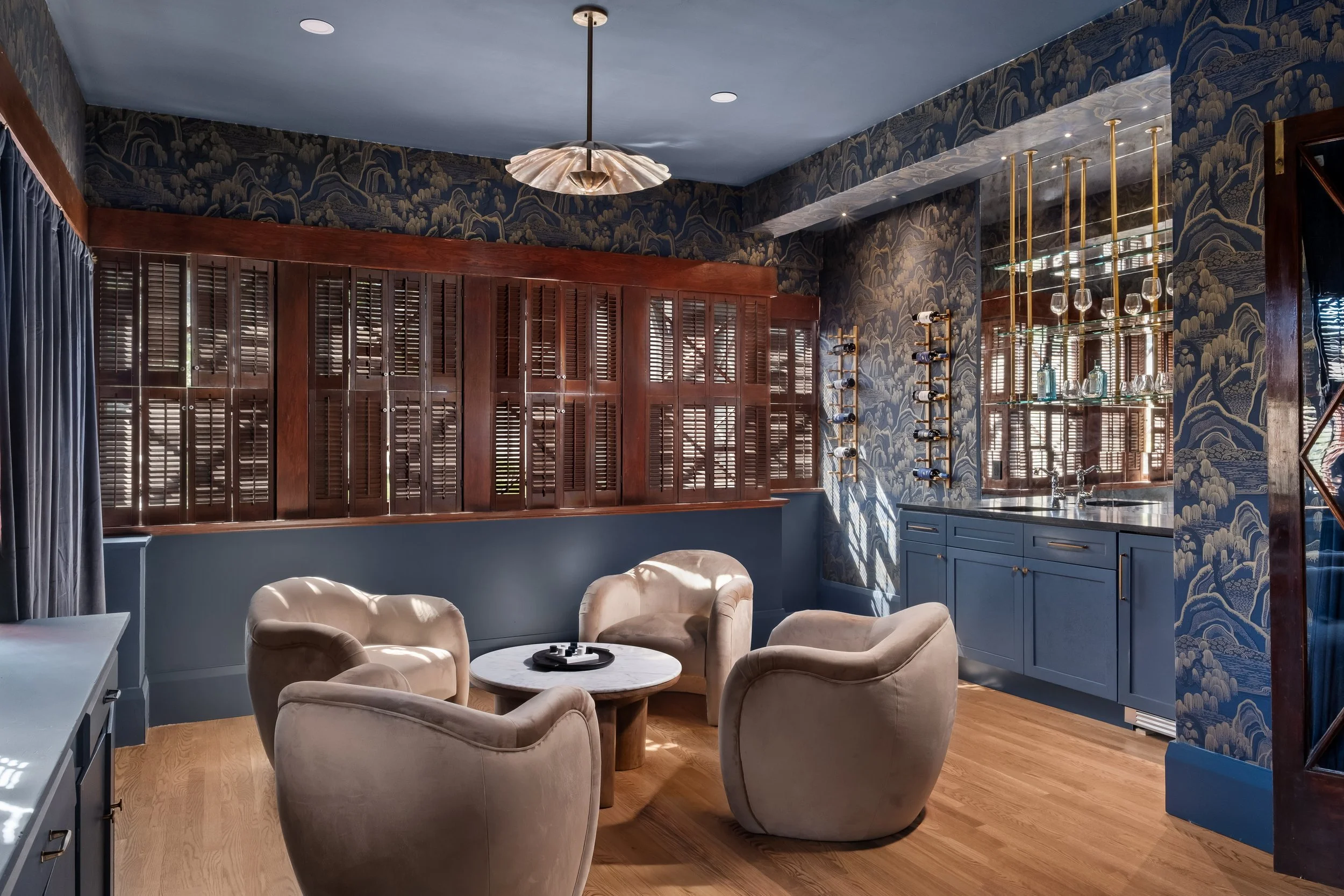 A cozy room with a blue wallpaper featuring mountain and cloud patterns, a wooden cabinet with shutters, a blue kitchen counter with gold hardware, a wine rack, and an area with beige armchairs and a round coffee table.