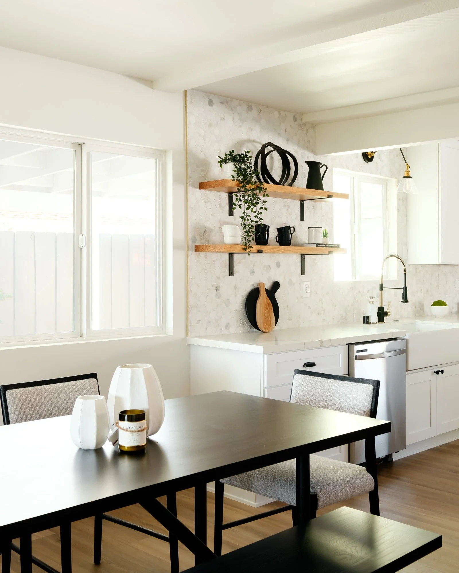 A modern kitchen and dining area with black table, cream chairs, shelves with black and white decorative items, and large windows letting in natural light.