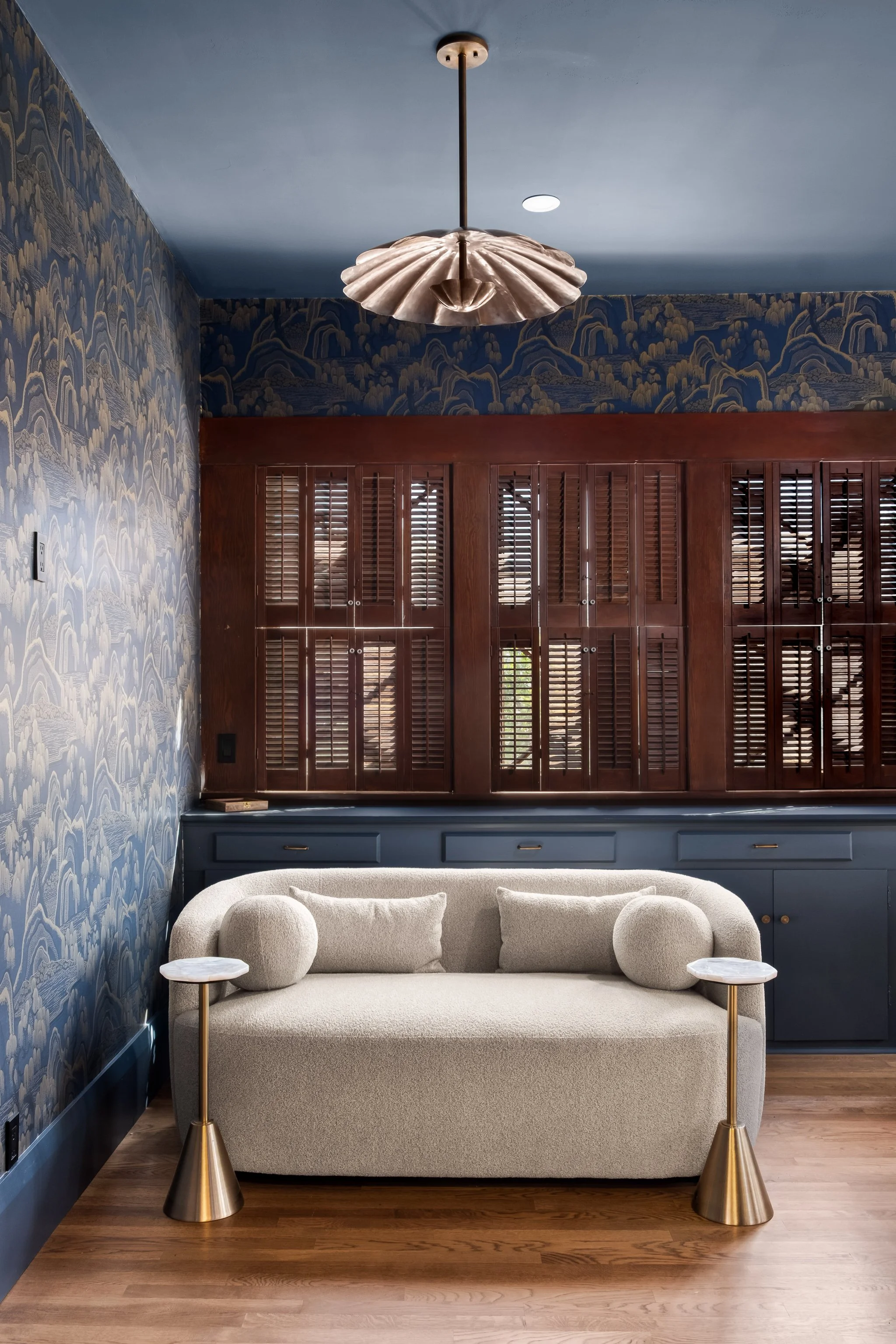 A living room with a cream-colored sofa, two side tables with gold bases, a wooden cabinet with shutters behind the sofa, patterned blue wallpaper on the walls, a light blue ceiling with a large decorative ceiling light, and hardwood floors.
