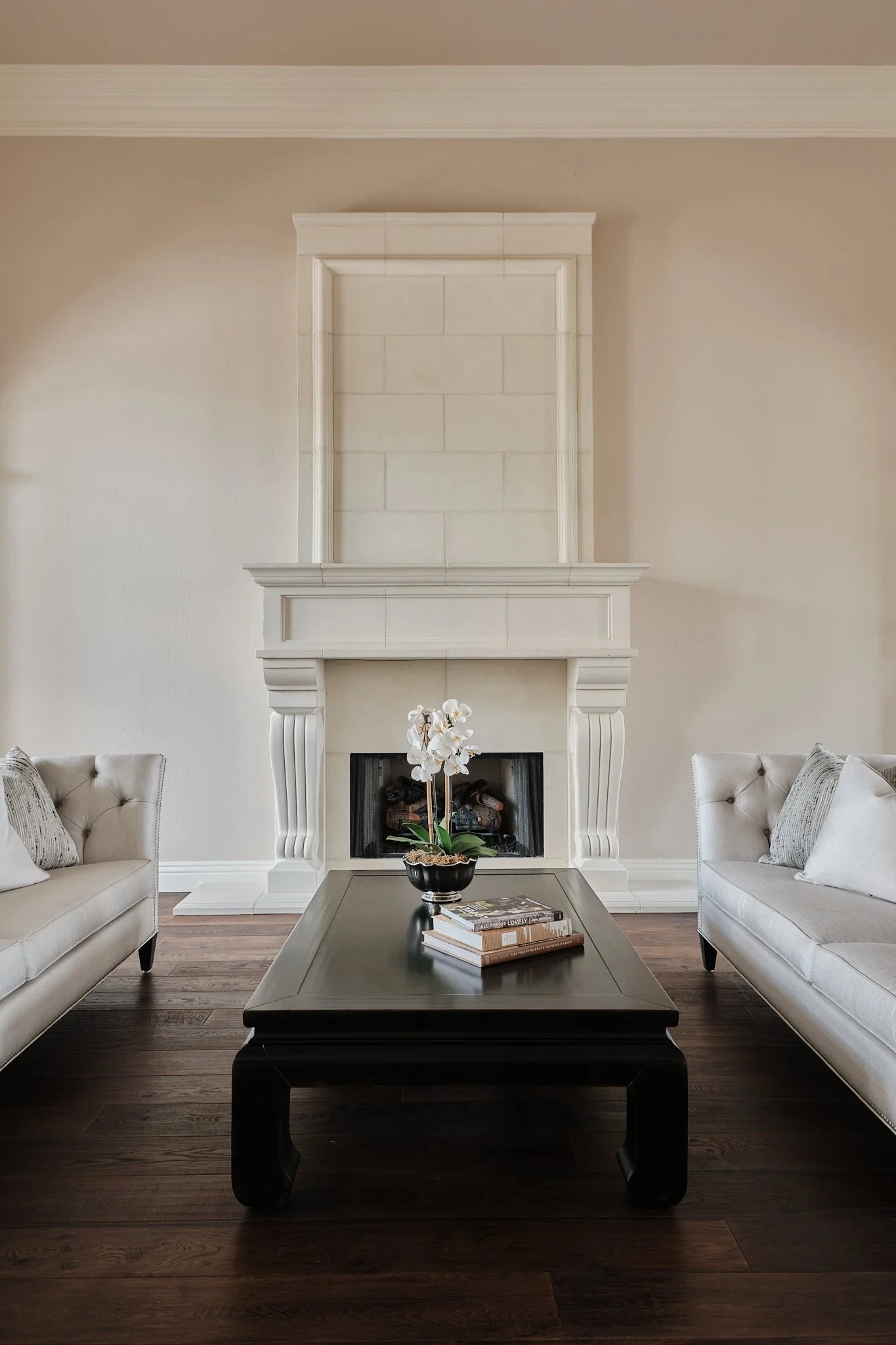 Living room with fireplace, two white sofas, a dark wooden coffee table with a potted orchid and books, and hardwood floors.