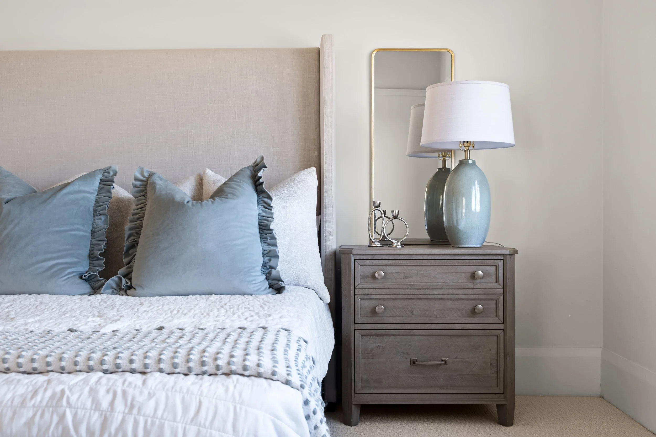A bedroom interior featuring a bed with multiple light blue and white pillows, a wooden nightstand with two drawers, a modern lamp with a white shade and ceramic base, and a tall rectangular mirror with a gold frame on the wall.