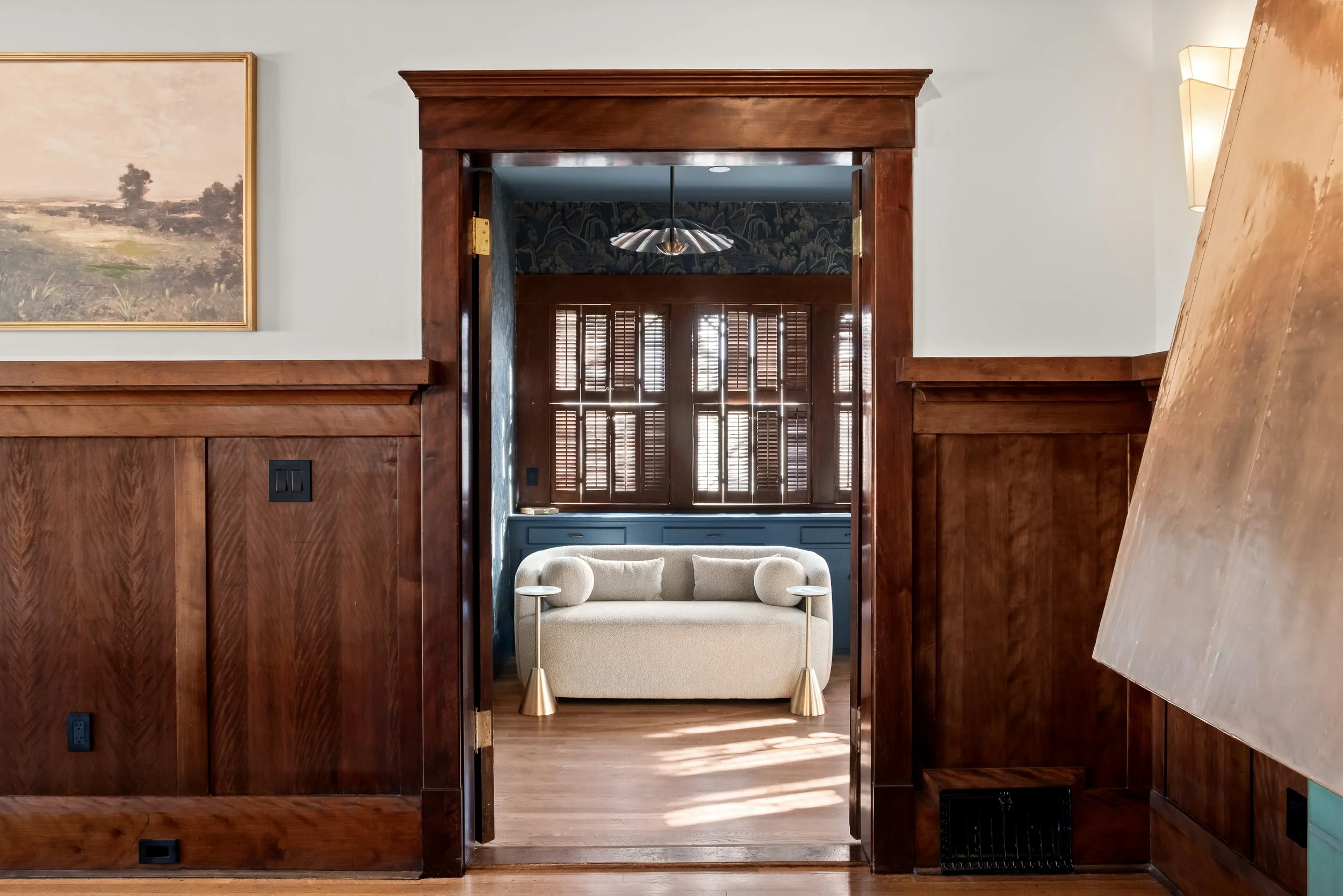 A view through a doorway showing a room with wooden wainscoting, a beige sofa, and large windows with wooden shutters.