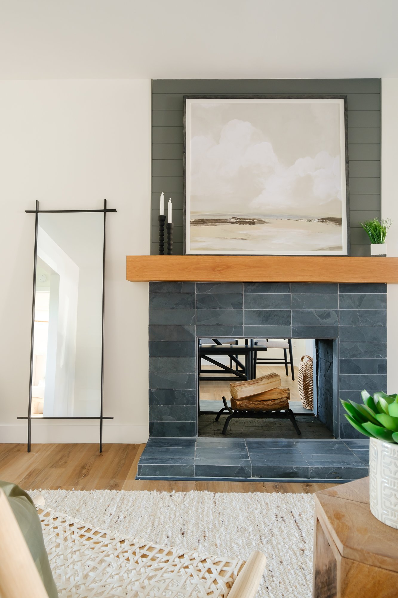 Living room with a fireplace, large abstract seascape painting above the mantel, black candlesticks, green plant in a pot, and modern decor, with a ladder-style mirror on the wall and a woven chair nearby.