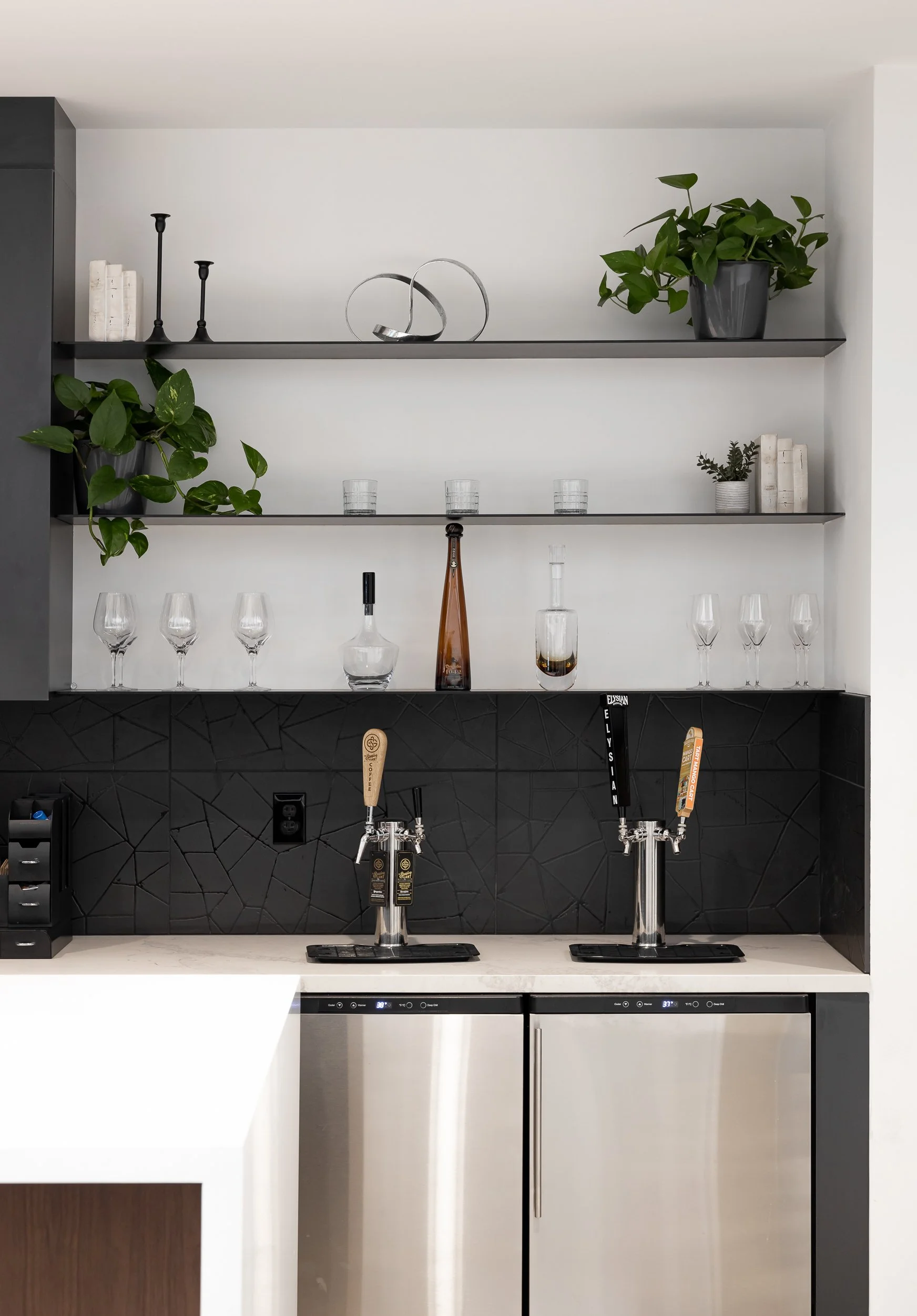 Modern bar area with black shelves holding decorative items, green plants, and books. Two beer tap dispensers on a white countertop below the shelves, and a stainless steel mini fridge underneath.