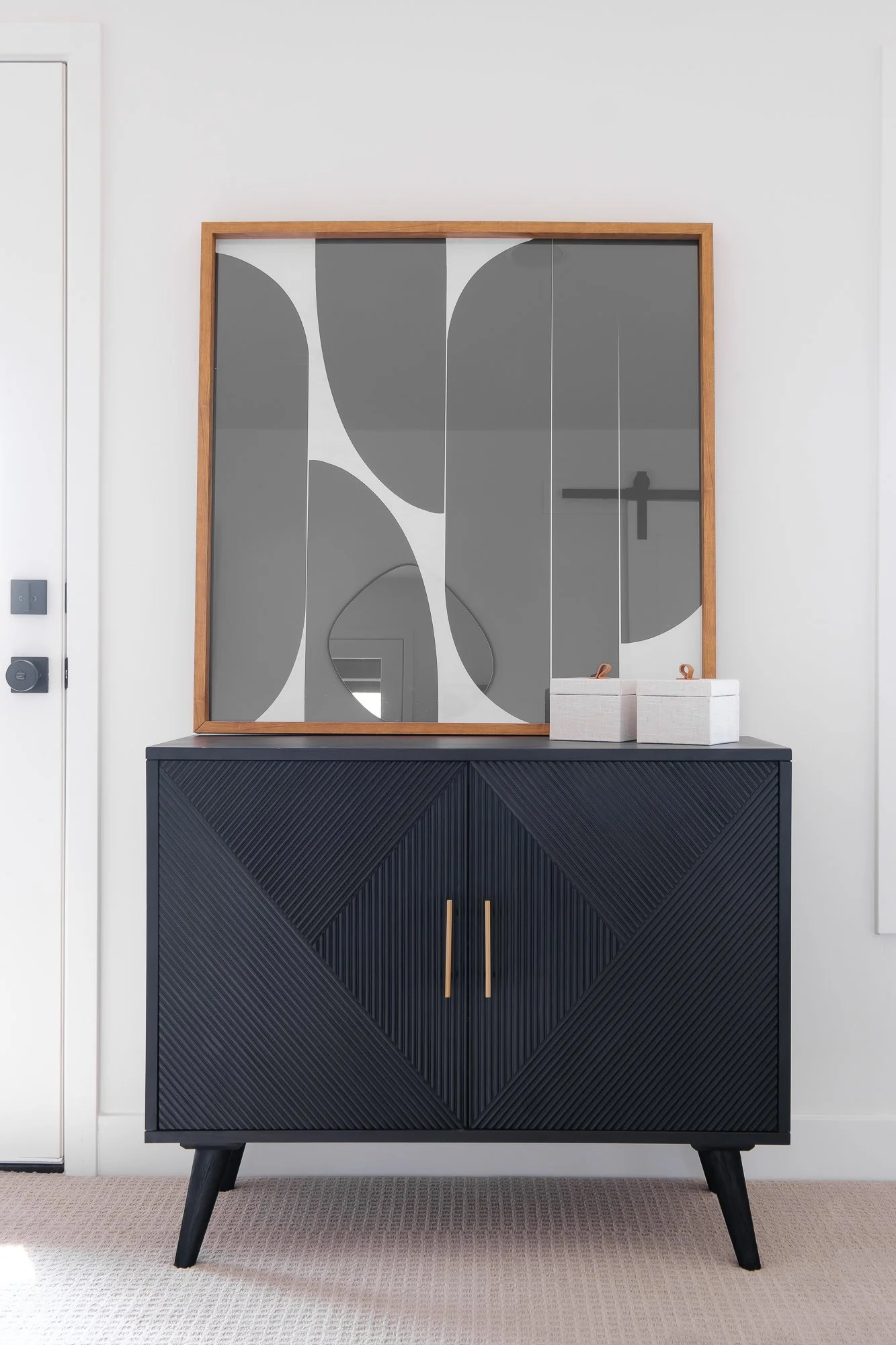 A black mid-century modern cabinet with a textured geometric pattern on its doors, standing on angled legs. On top, there are two white boxes and a reflective mirror with a wooden frame hanging above it on a white wall.