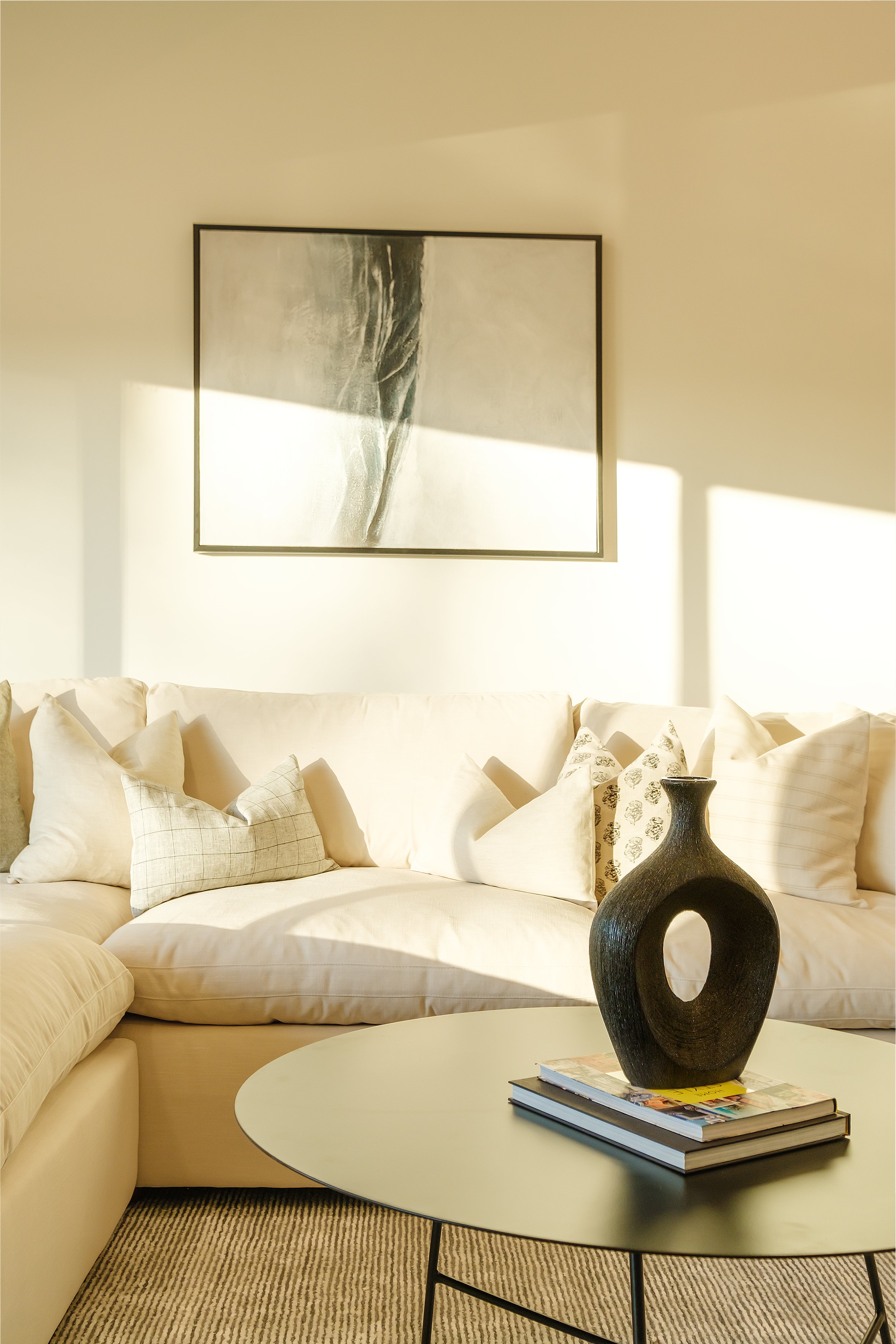 Living room with cream-colored sofa, decorative pillows, abstract artwork on the wall, black vase on a coffee table, and sunlight streaming in.