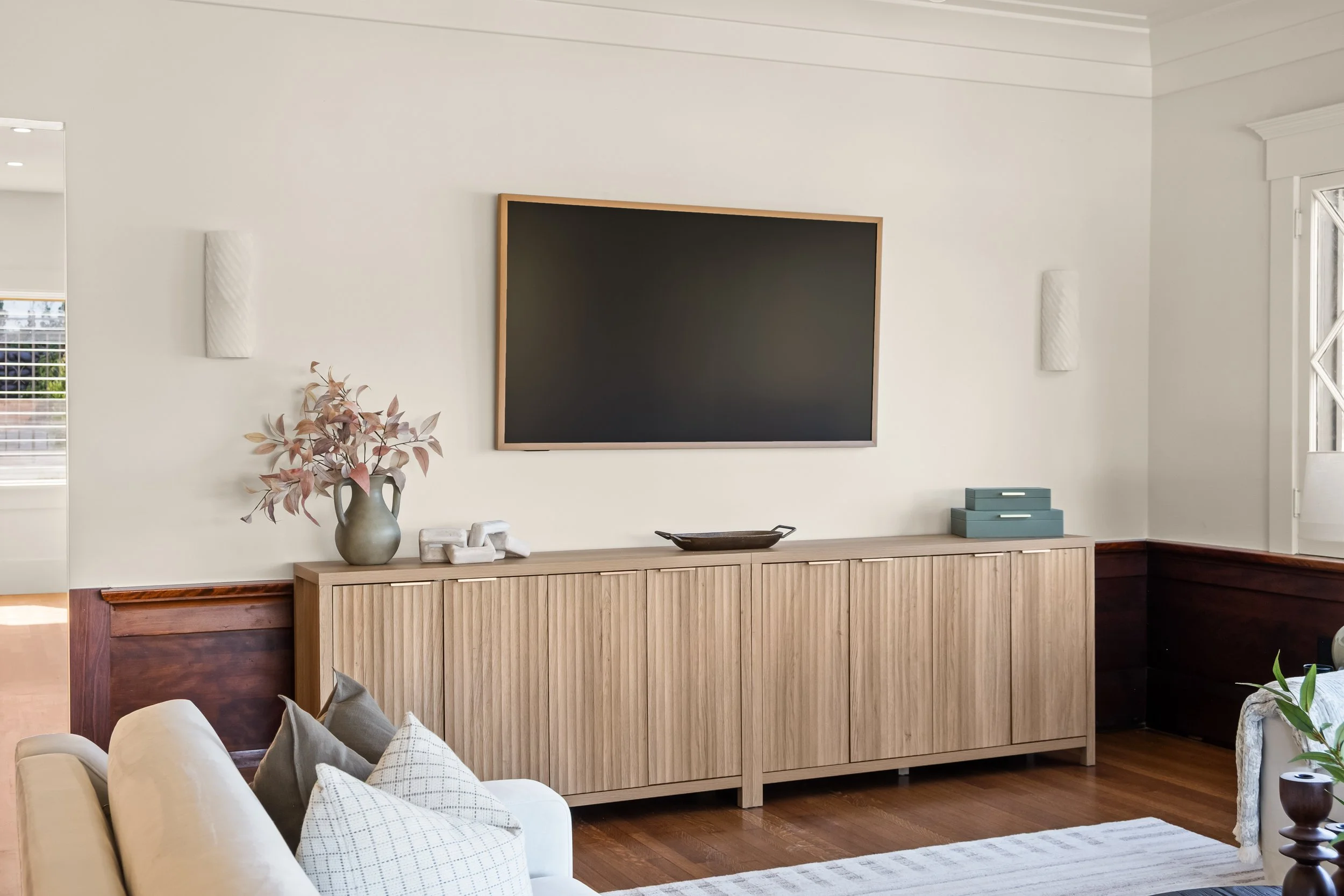 Living room with a wall-mounted flat-screen TV, decor items including a vase with dried leaves, a decorative sculpture, and a low wooden media console. Part of a white sofa with pillows is visible in the foreground, and there is a window with white t