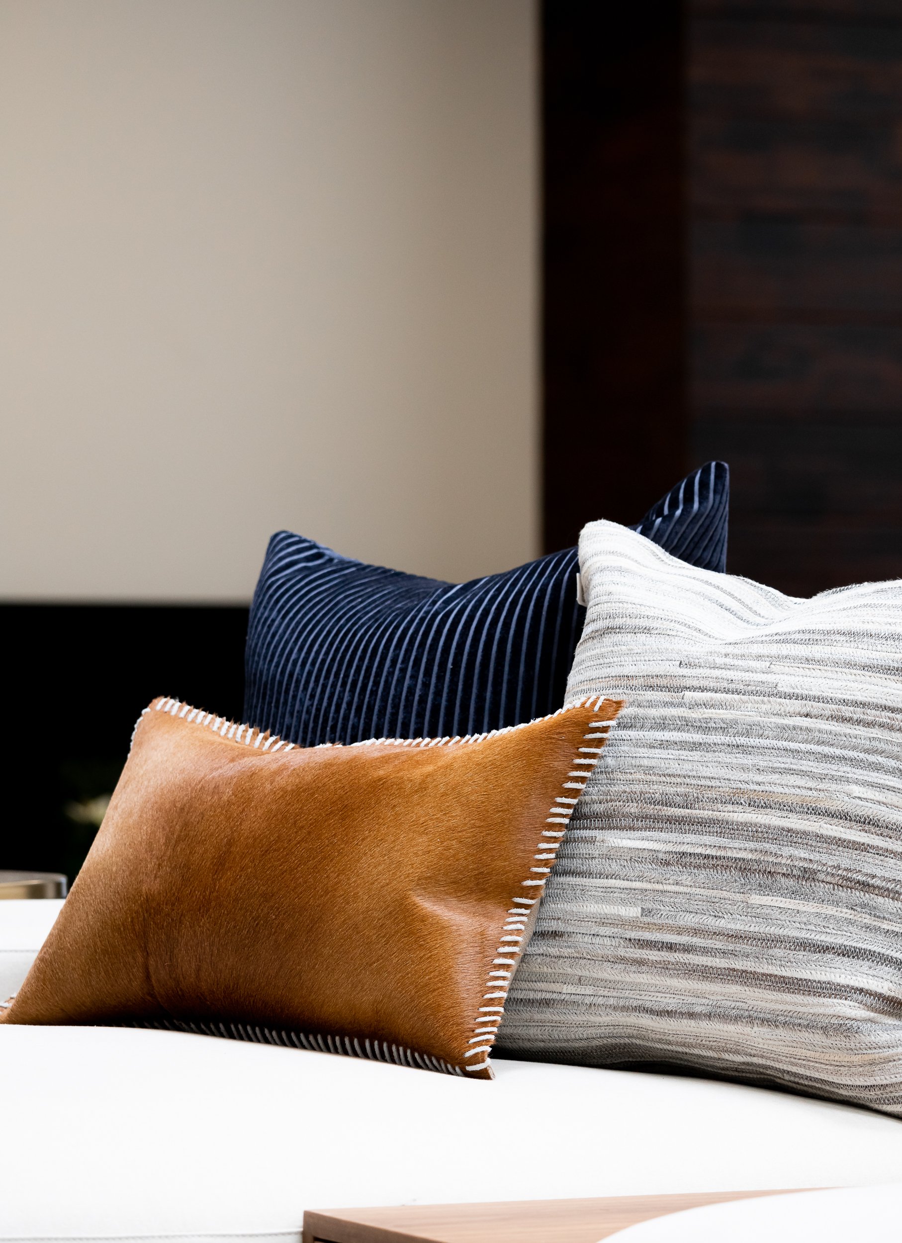 Close-up of decorative pillows arranged on a sofa, including a tan pillow, a navy blue pillow with white pinstripes, and a white pillow with gray stripes, in a modern interior setting.