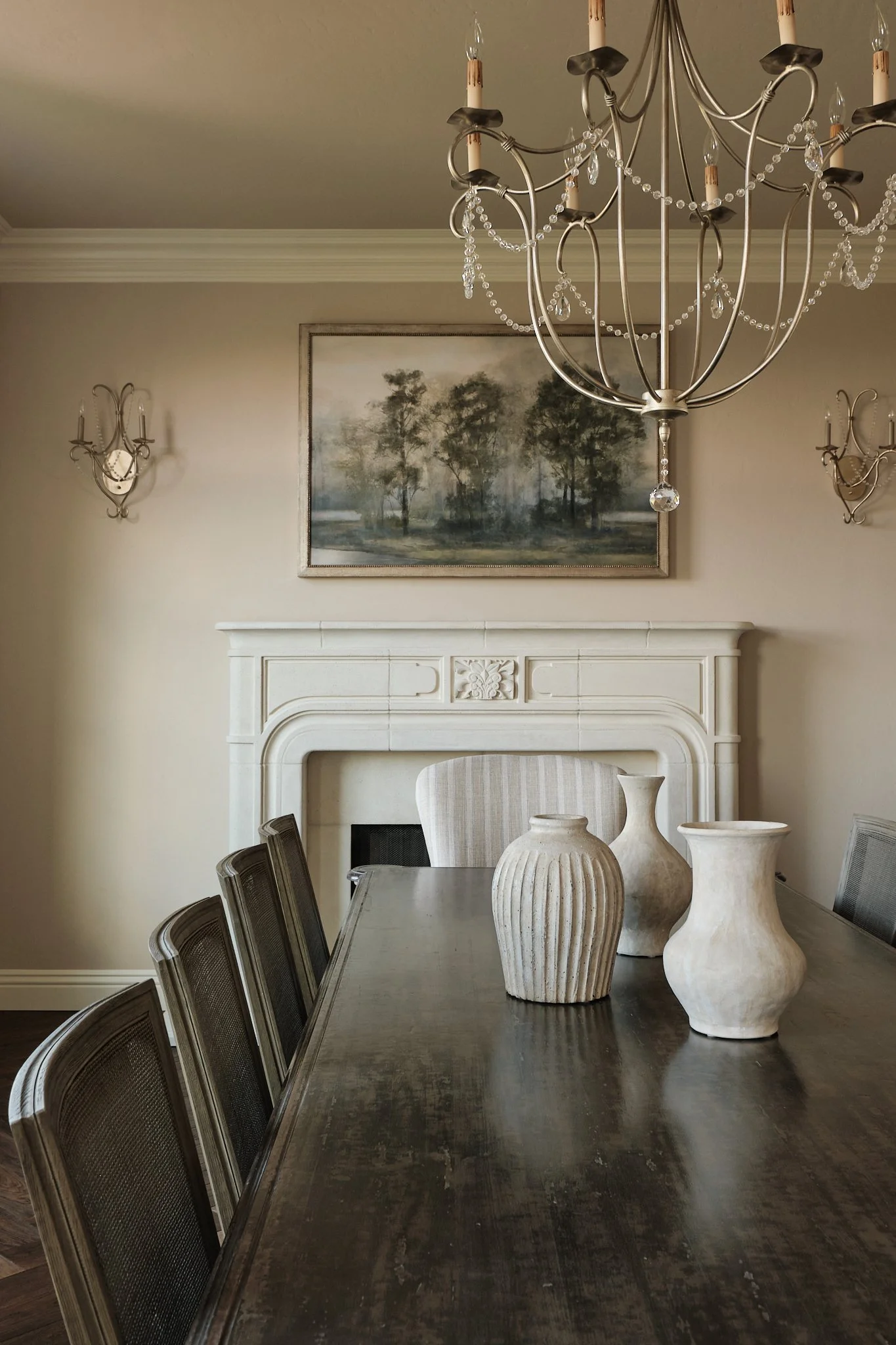 A dining room with a dark wooden table, surrounded by several chairs with mesh backs. On the table, there are five large beige vases of various shapes and sizes. The background features a white fireplace mantle, a landscape painting of trees on the w