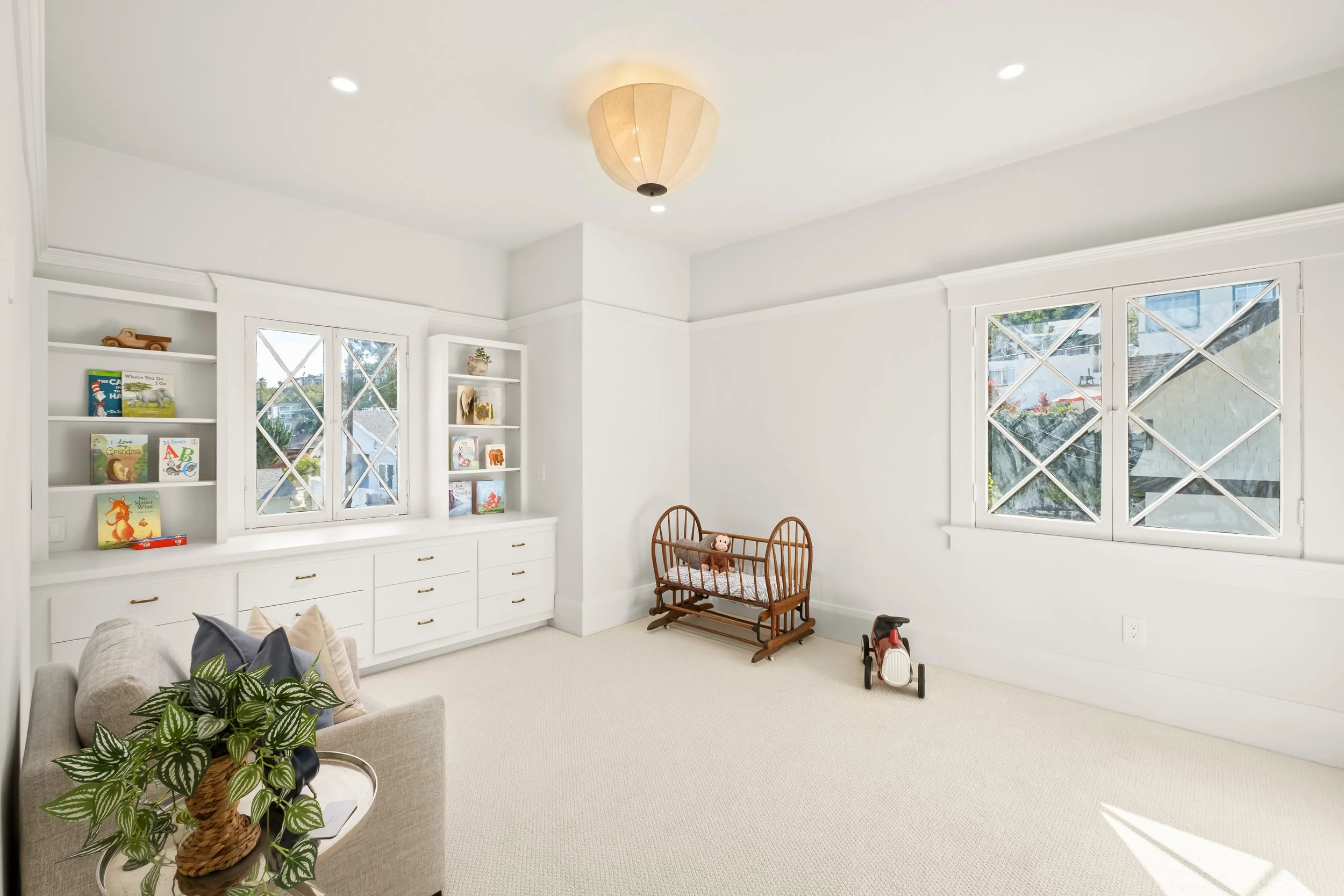 Bright, white living room with large windows, a built-in bookshelf with children’s books, a wooden crib, some toys, a small couch with pillows, a potted plant, and a modern ceiling light