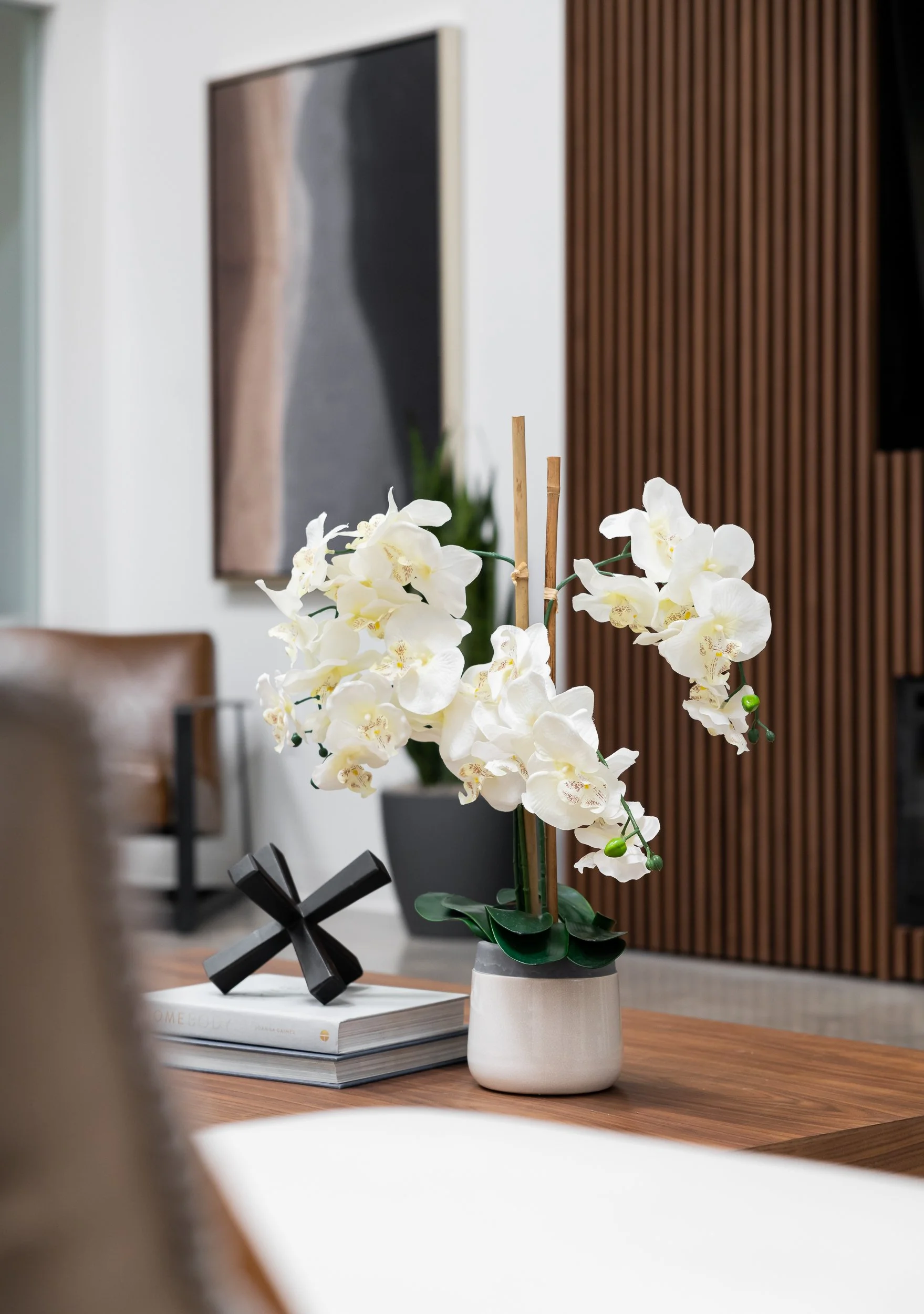 A white orchid plant in a white ceramic pot on a wooden table, with a black geometric sculpture and a stack of books nearby, in a modern interior setting.