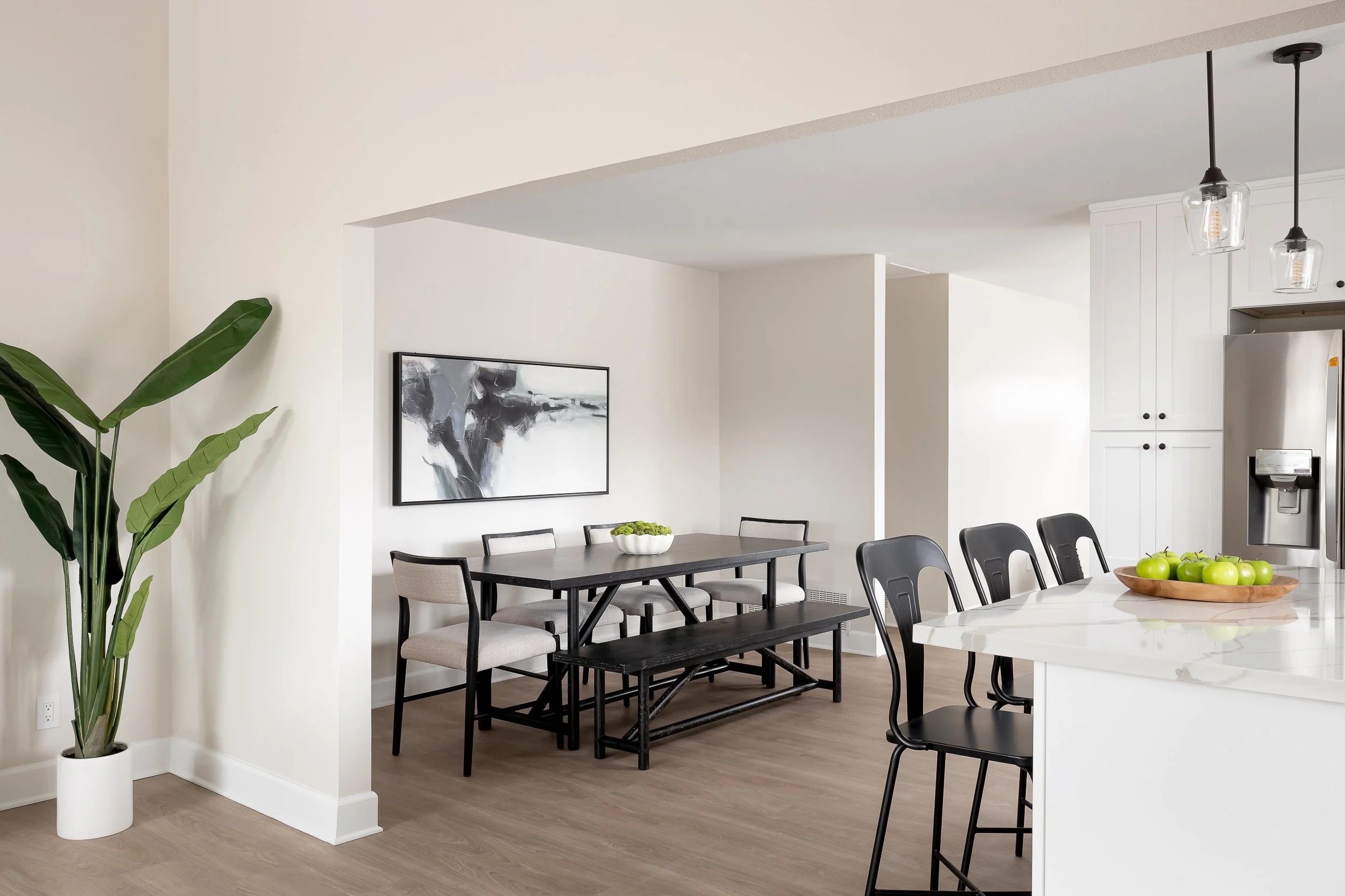 A modern dining area with a large white marble table, black chairs, and a bowl of green apples. The room has white walls, a painting, large green plant in a white pot, and hanging light fixtures.