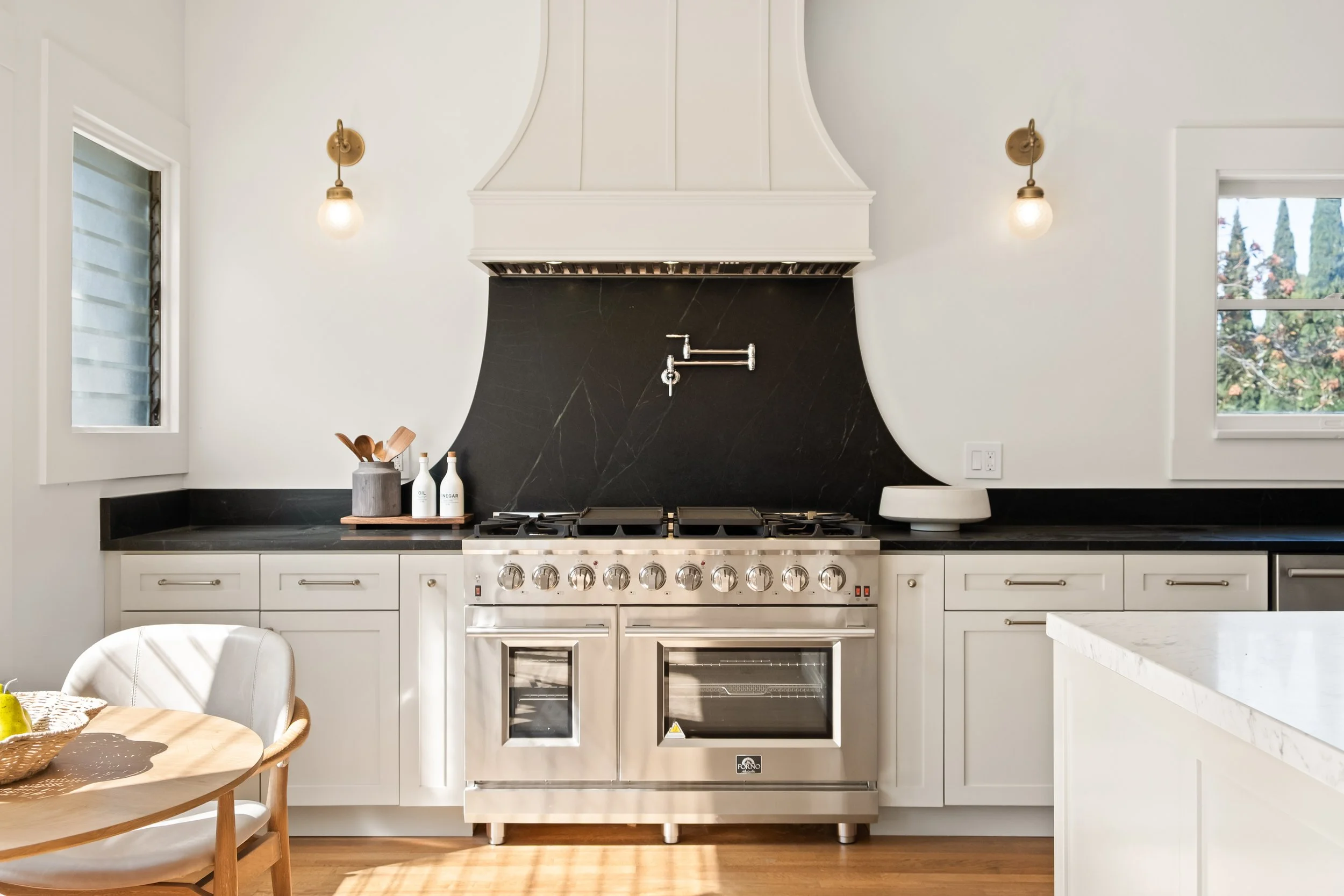 Modern kitchen with white cabinetry, black countertops, and a stainless steel stove and oven. There are also two small windows and decorative items on the counter.