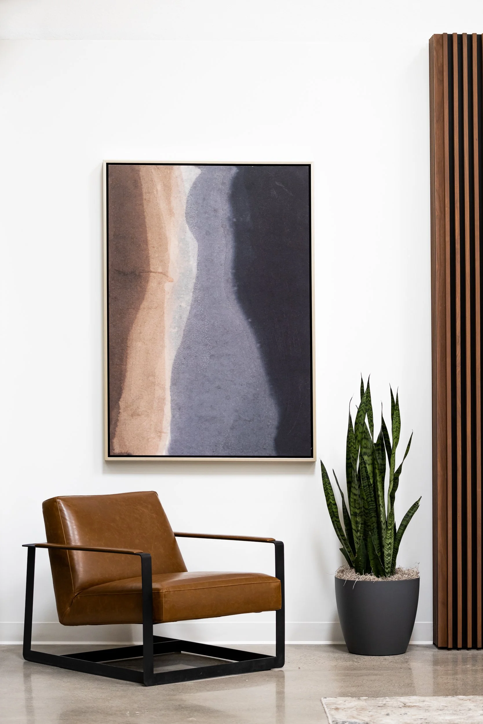 A modern living room corner with a brown leather armchair, a large abstract landscape painting on the white wall, a tall snake plant in a gray pot, and a wooden slatted room divider to the side.