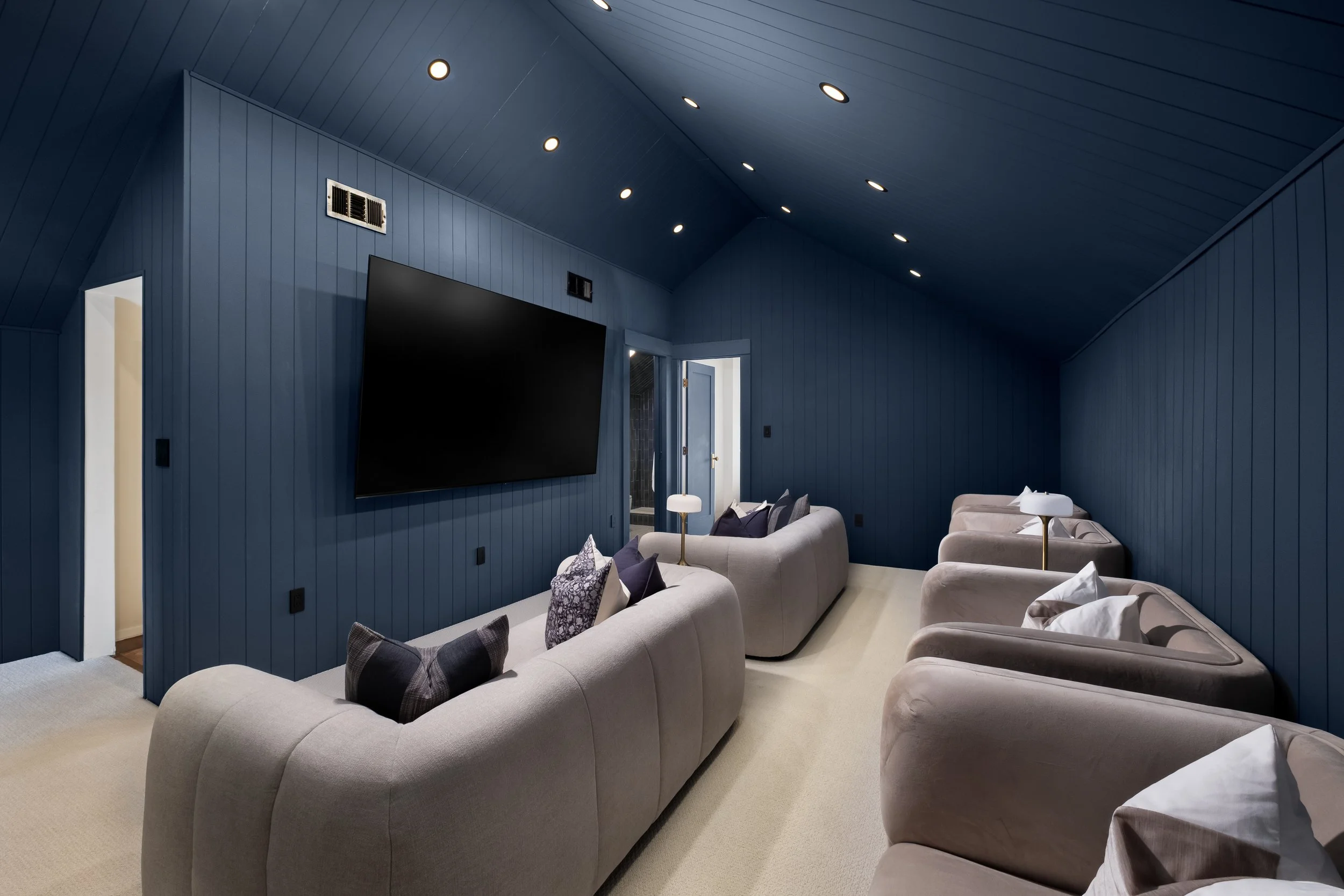 A cozy living room with blue paneled walls and ceiling, featuring recessed lighting, a flat screen TV, and multiple light-colored sofas with cushions.