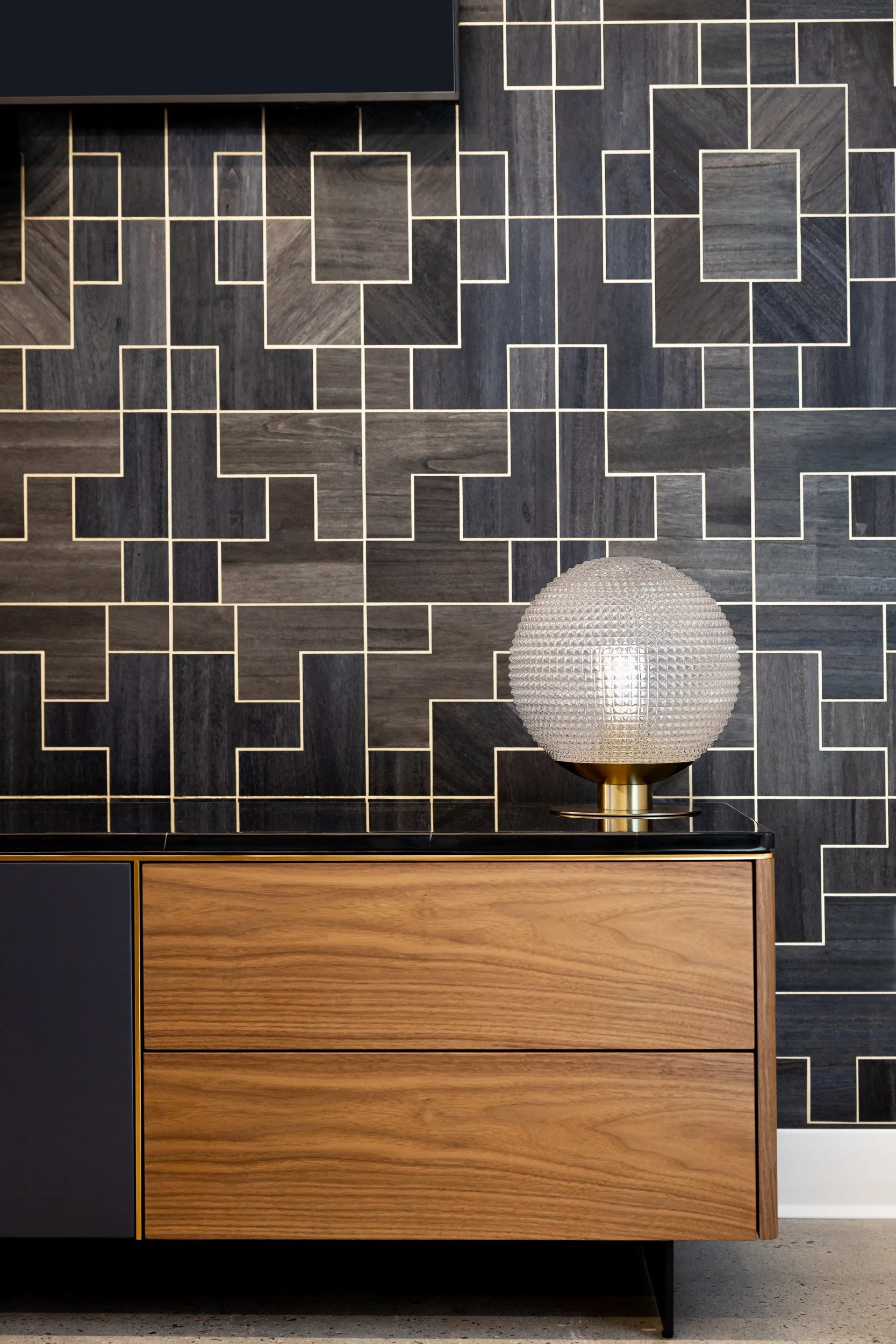 Decorative black and gray tiled wall with wooden cabinet and a spherical textured lamp with a brass base on top.
