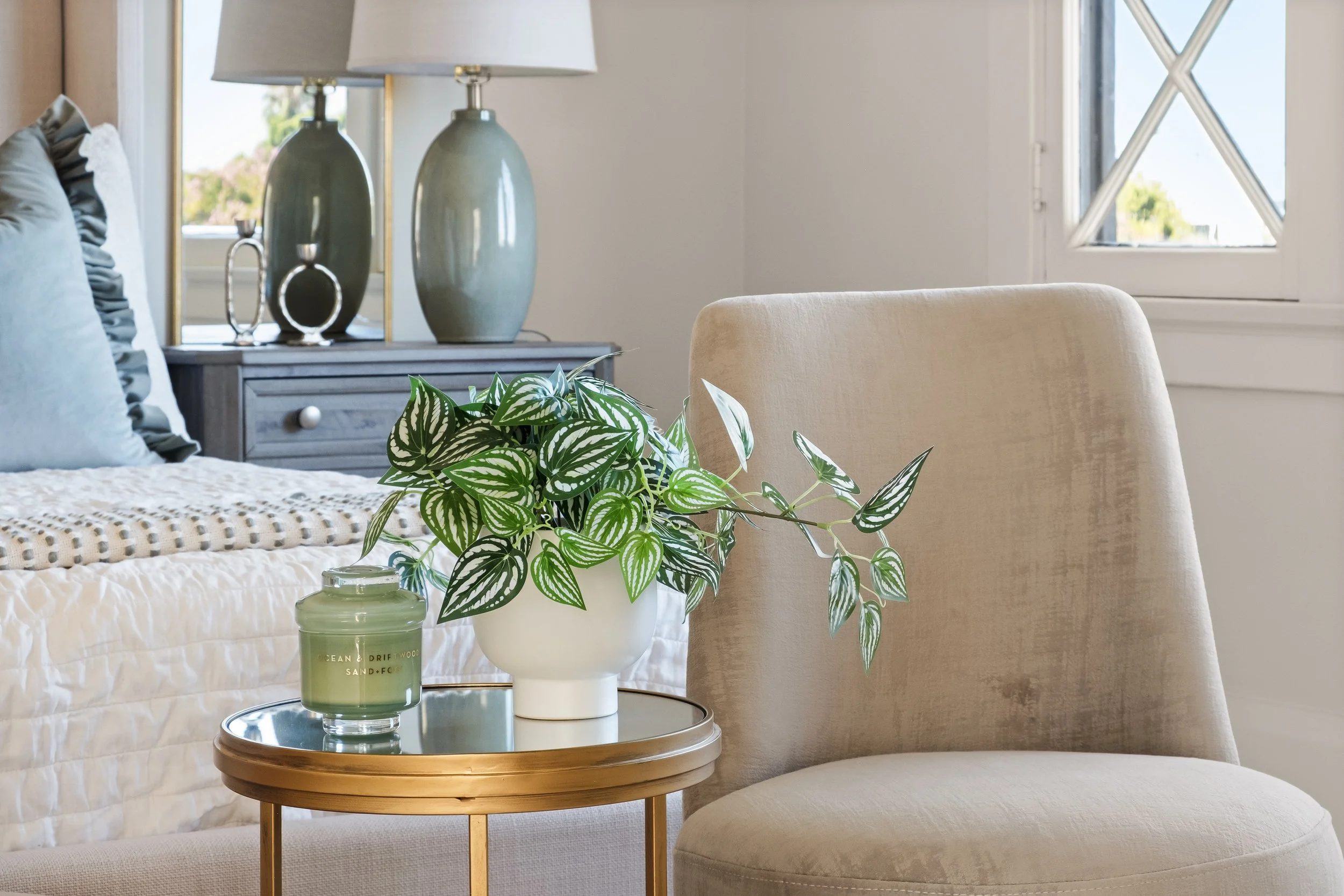 A cozy bedroom corner with a white bed, decorative pillows, a nightstand with two green vases and a silver sculpture, a table with green candles and a potted plant, beige upholstered armchair, and a window with a view of greenery outside.