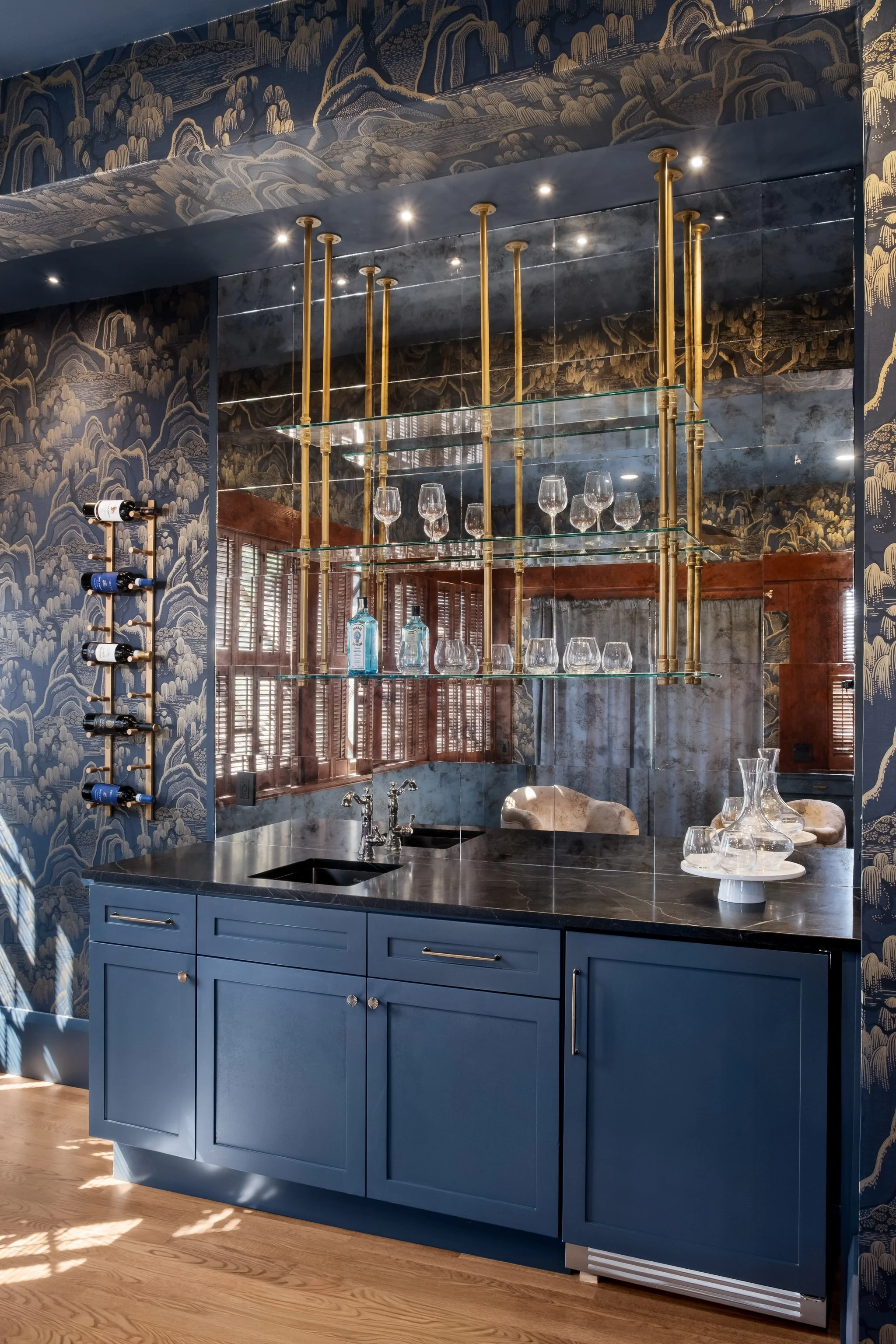 A kitchen with blue cabinets, black marble countertop, and brass shelving holding glassware. The wall behind has a dark patterned wallpaper with gold accents, and a window with wooden blinds. There is a wine rack on the side of the wall, and a sink w