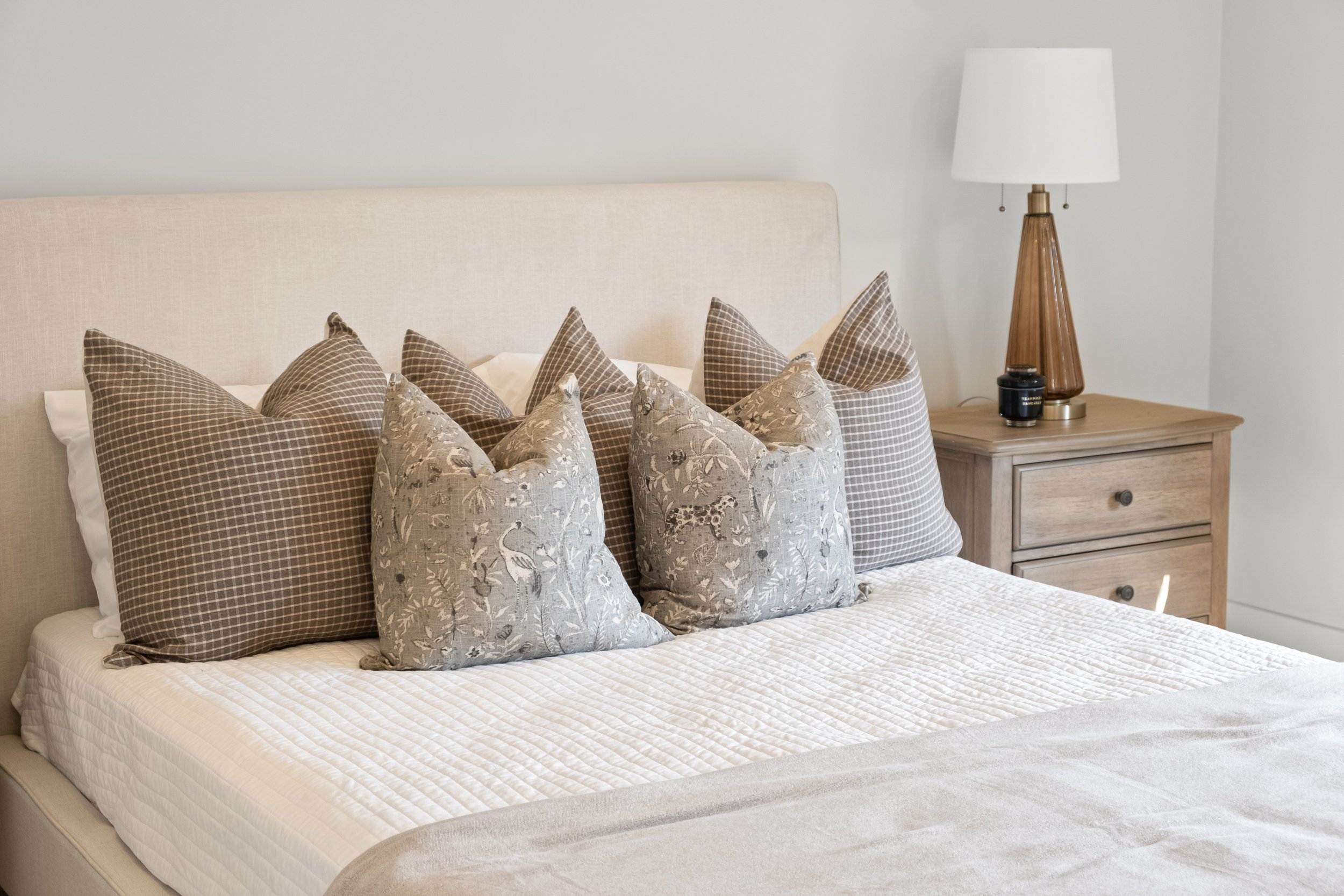 A neatly made bed with a beige upholstered headboard, beige and patterned throw pillows, and a white quilted bedspread. To the right, there is a wooden nightstand with a table lamp with a white lampshade and a wooden base, along with a black containe