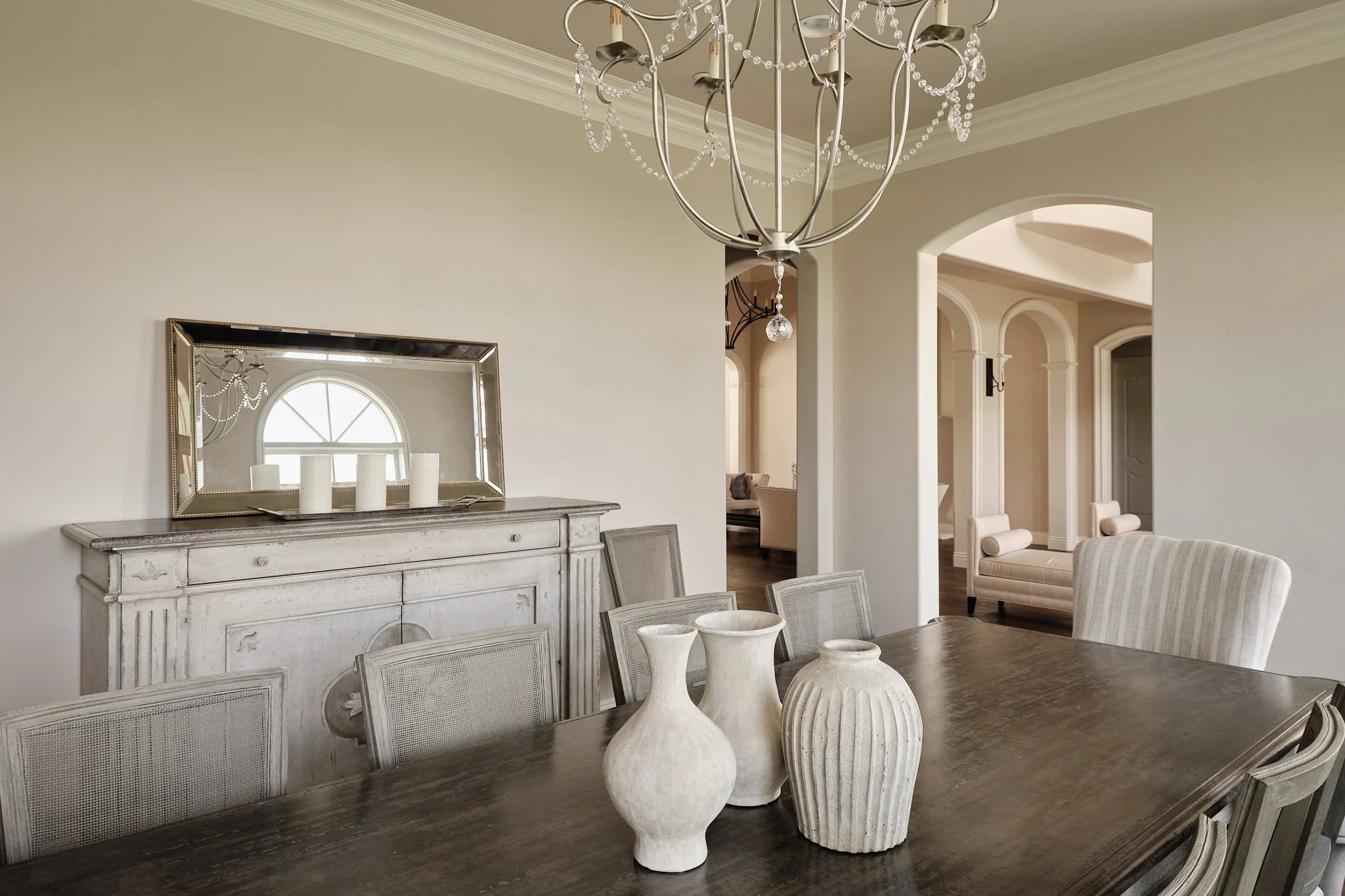Dining room with a large wooden table, ceramic vases, a distressed white sideboard with candles, a mirror reflecting a church-style window, and a chandelier with hanging crystals.