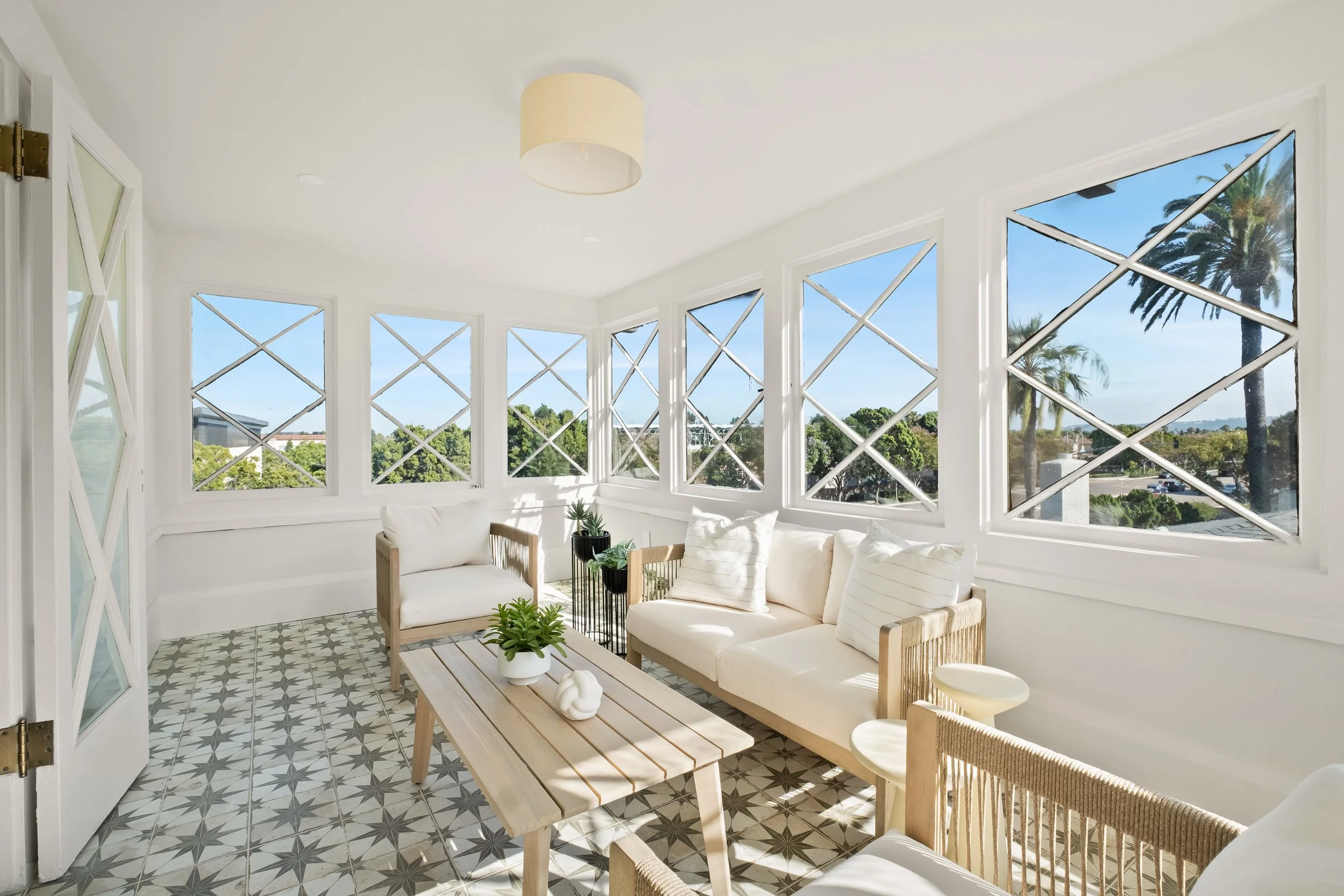 Bright sunlit enclosed porch with white walls and patterned tile floor, furnished with white cushioned sofas, wooden chairs, a wooden table with decorative items, and large windows showing a view of palm trees and a blue sky.