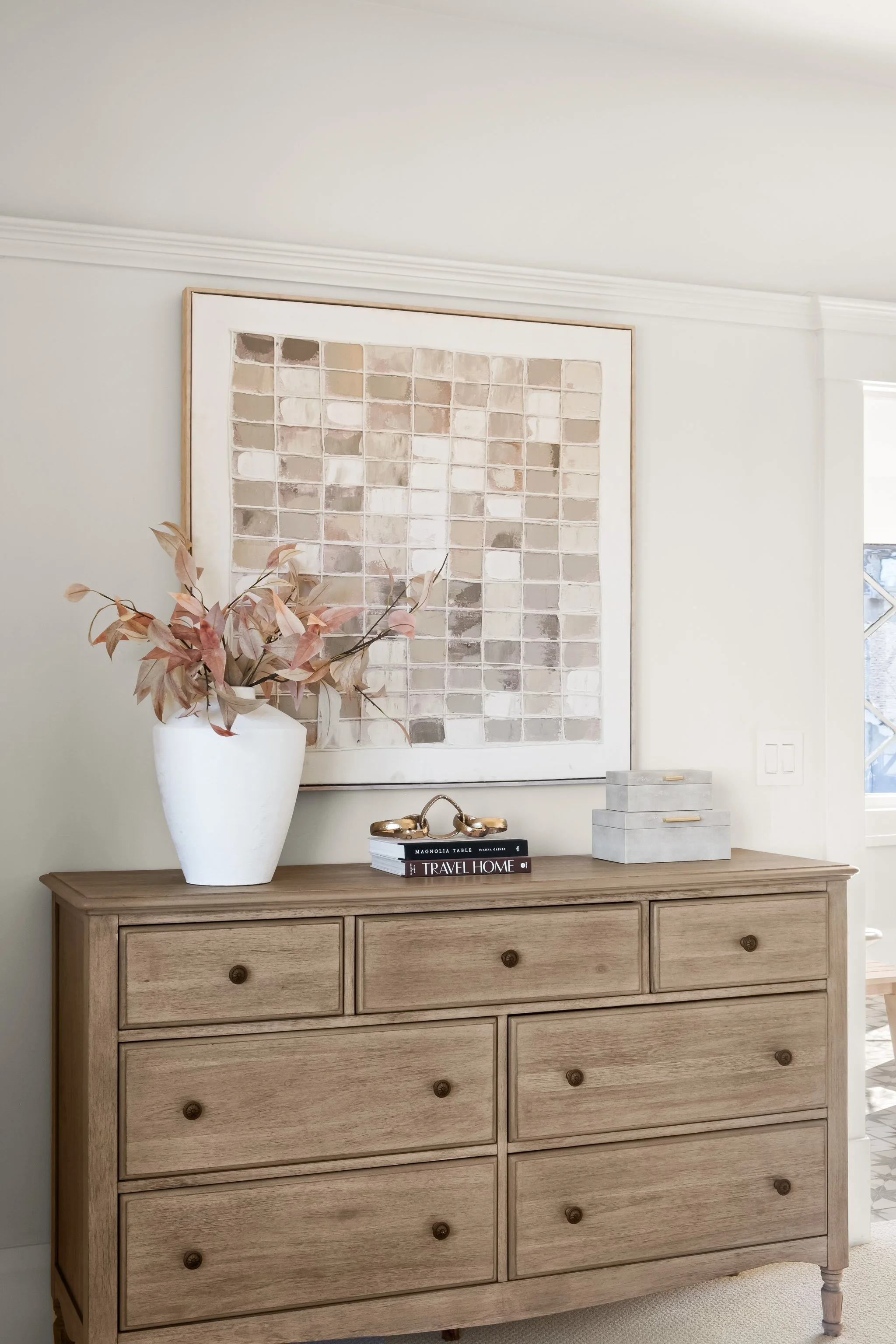 A wooden dresser with nine drawers, decorated with a large white vase holding dried pinkish leaves, a small stack of books titled 'Magnolia Table' and 'Travel Home,' a gold decorative object, and a pastel abstract art piece on the wall behind. There 