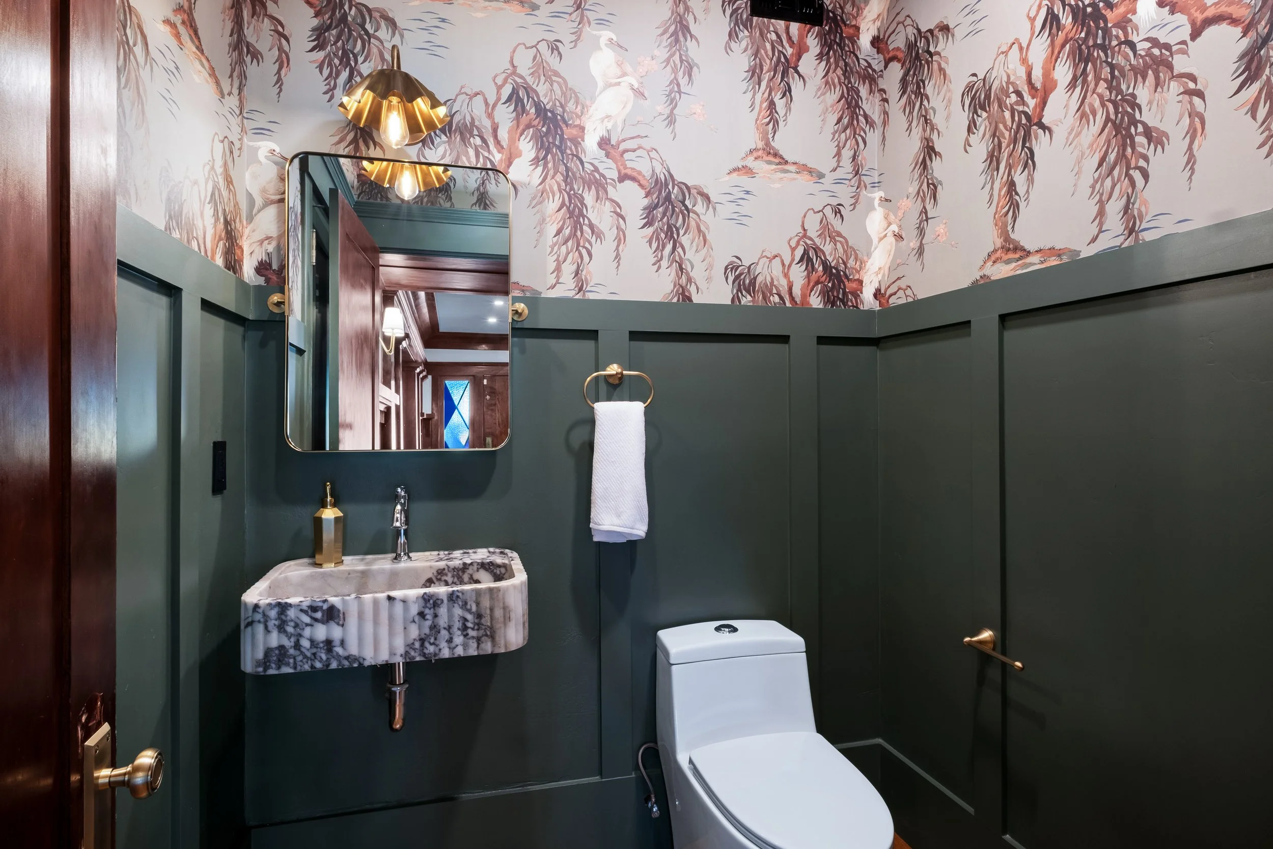 This is a bathroom with dark green wainscoting, a white toilet, a marble sink with a gold faucet, a rectangular mirror, a gold wall sconce with Edison style bulbs, and wallpaper with a design of white cranes and hanging willow branches.