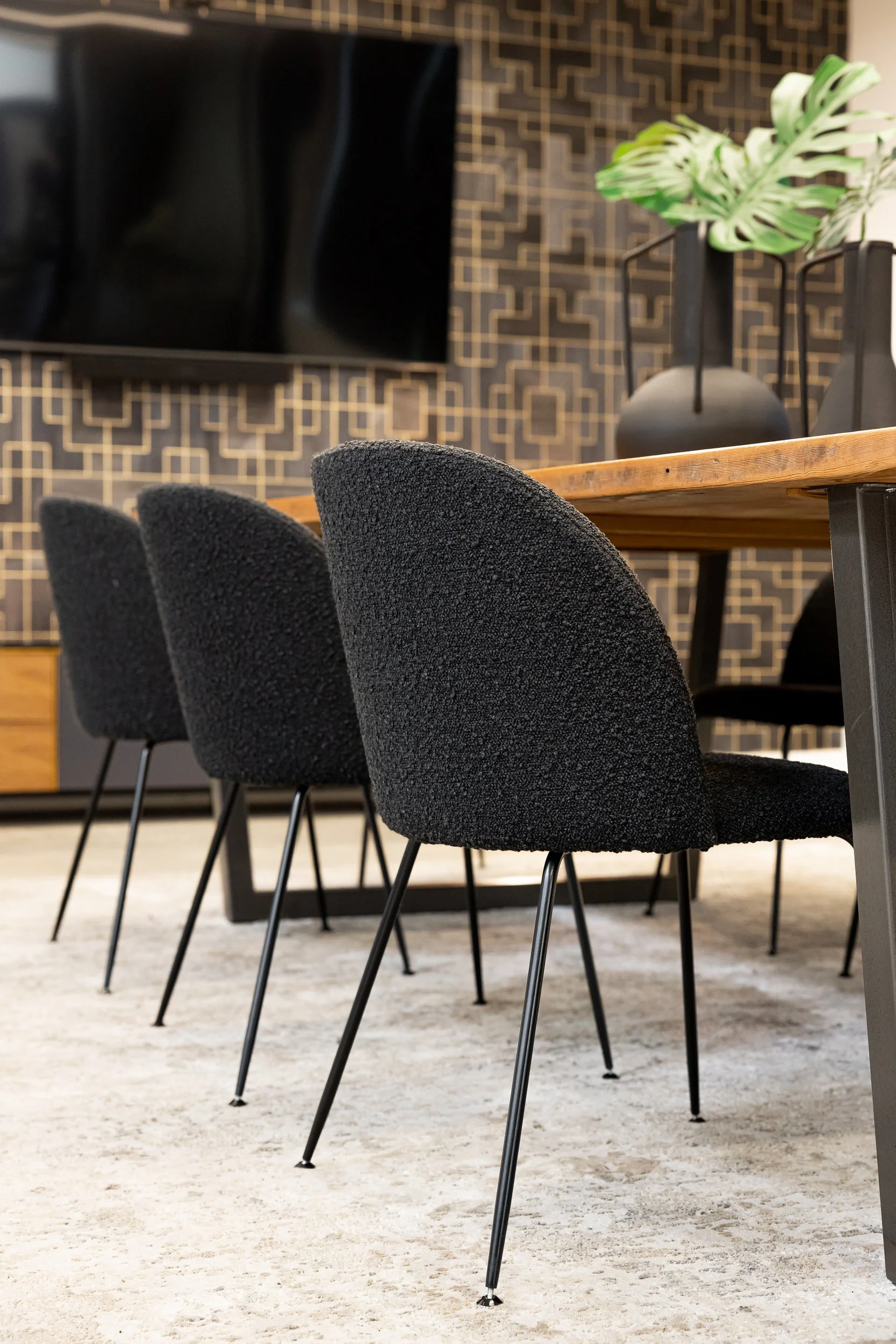 A modern dining area with a wooden table, black upholstered chairs on metal legs, a patterned black and gold accent wall, and a decorative plant in black vases.