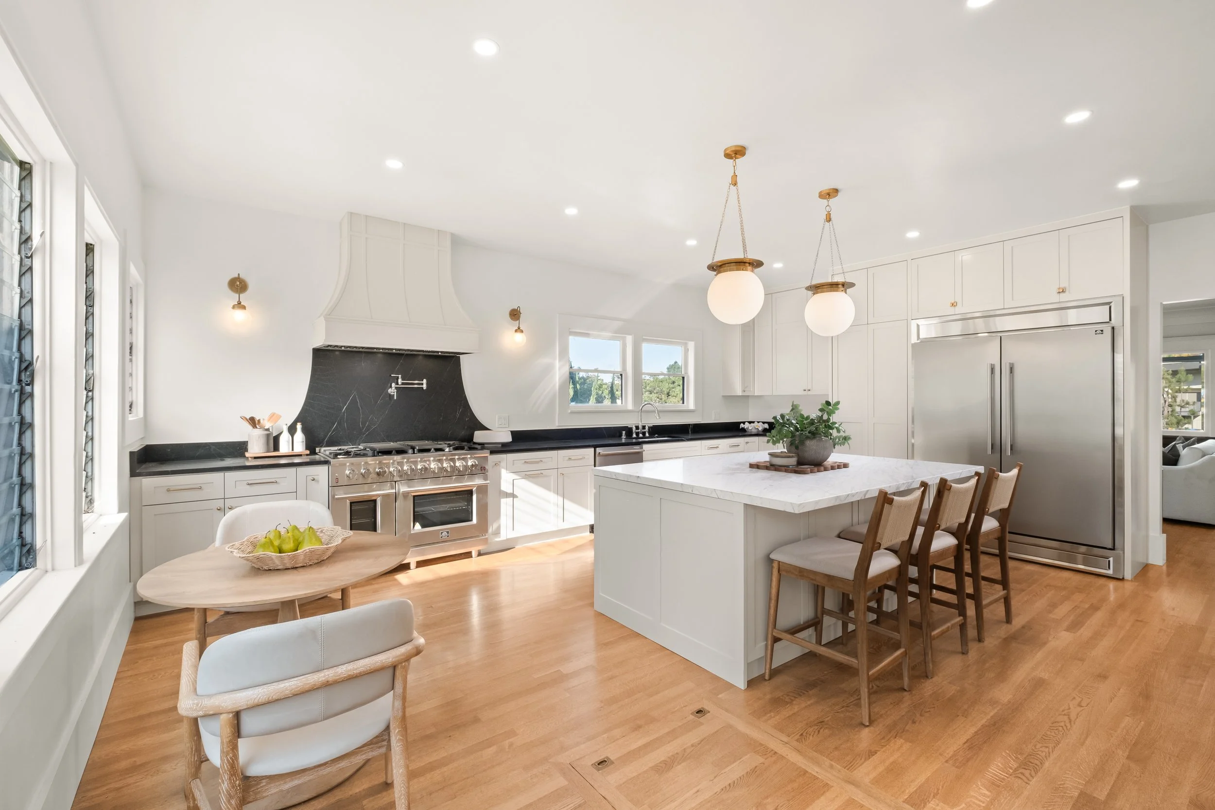 A modern, bright kitchen with white cabinets, a black marble backsplash, a stainless steel range, and a kitchen island with a white marble countertop. There are two pendant lights hanging above the island, and a basket of green apples on a wooden rou