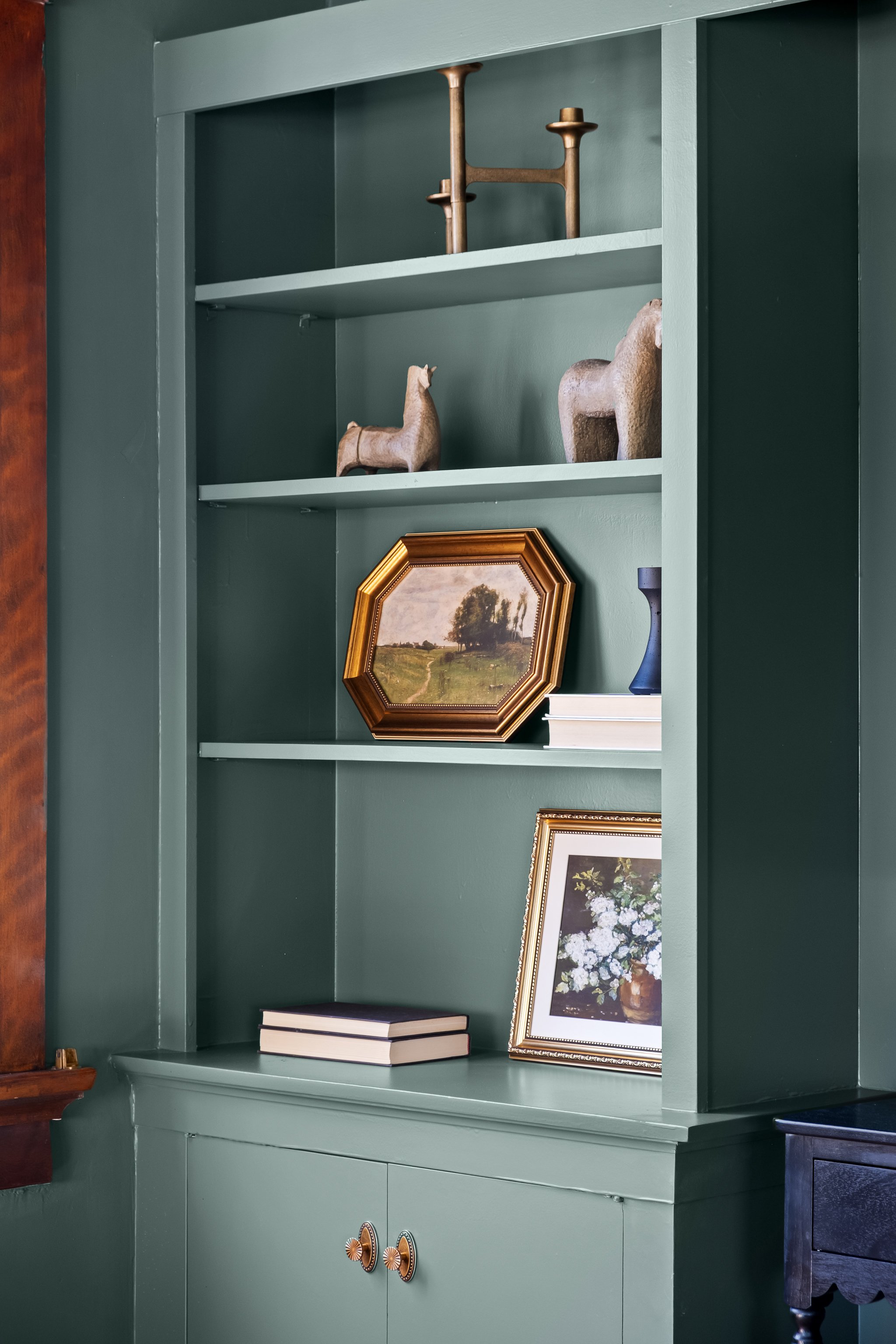 Green bookshelf with decorative objects, framed artwork, books, and a small blue vase.