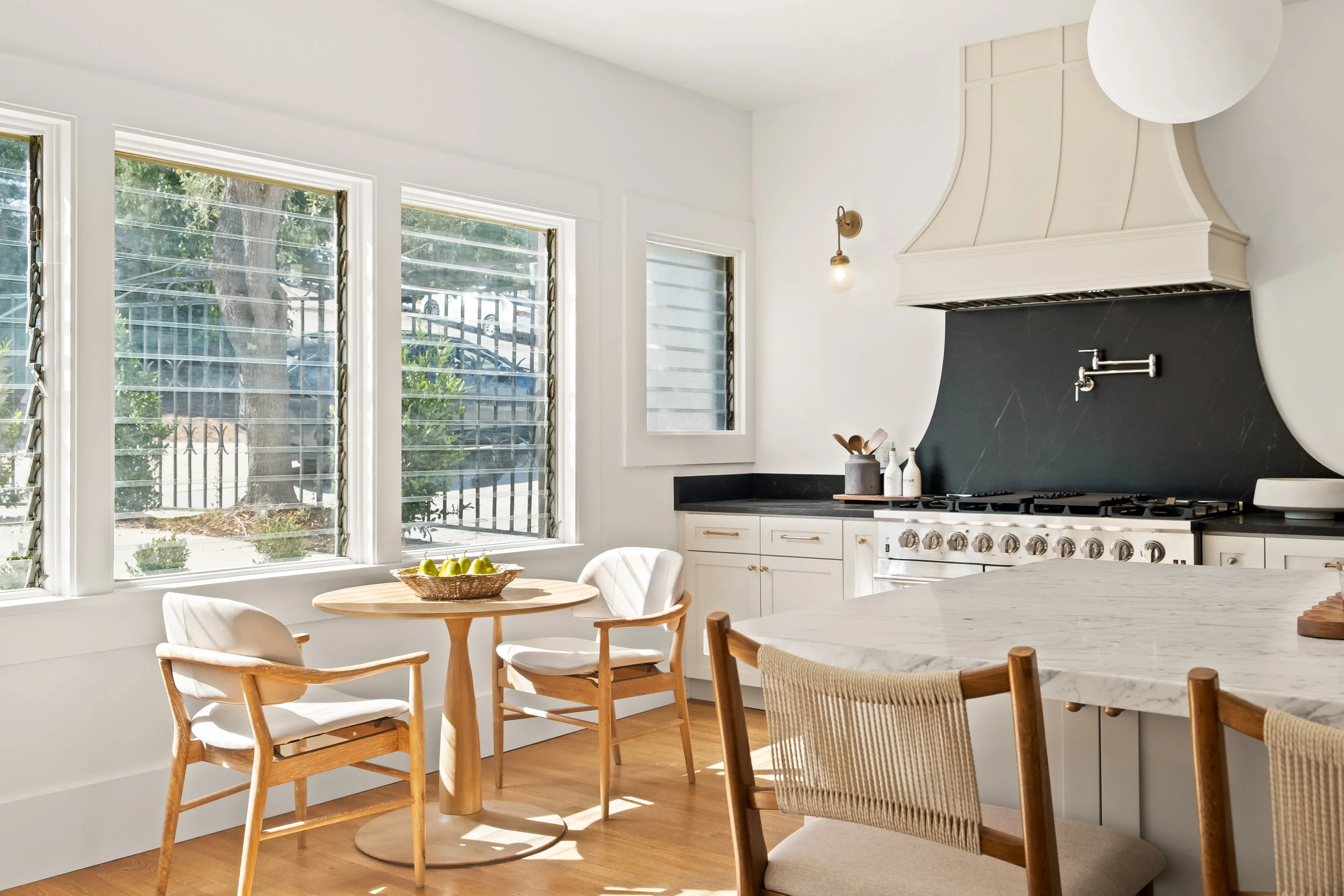Bright and airy kitchen with large windows, white cabinetry, black countertops, a white range hood, and wooden chairs around a marble dining table. A bowl of green apples is on the table, and sunlight streams through the windows.