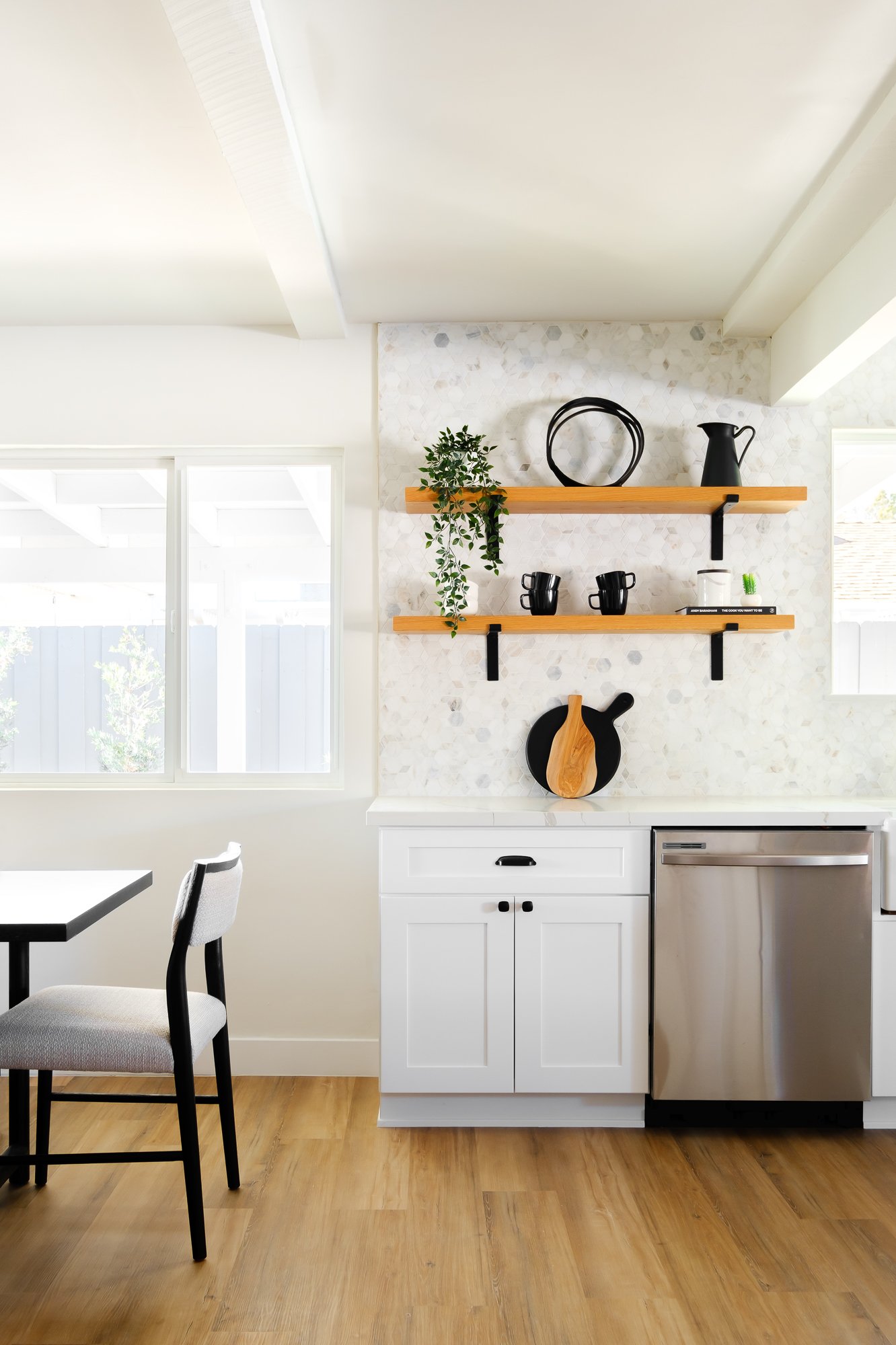 A modern kitchen with white cabinets, wooden shelves, and black and wood utensils, featuring a large window and wooden flooring.