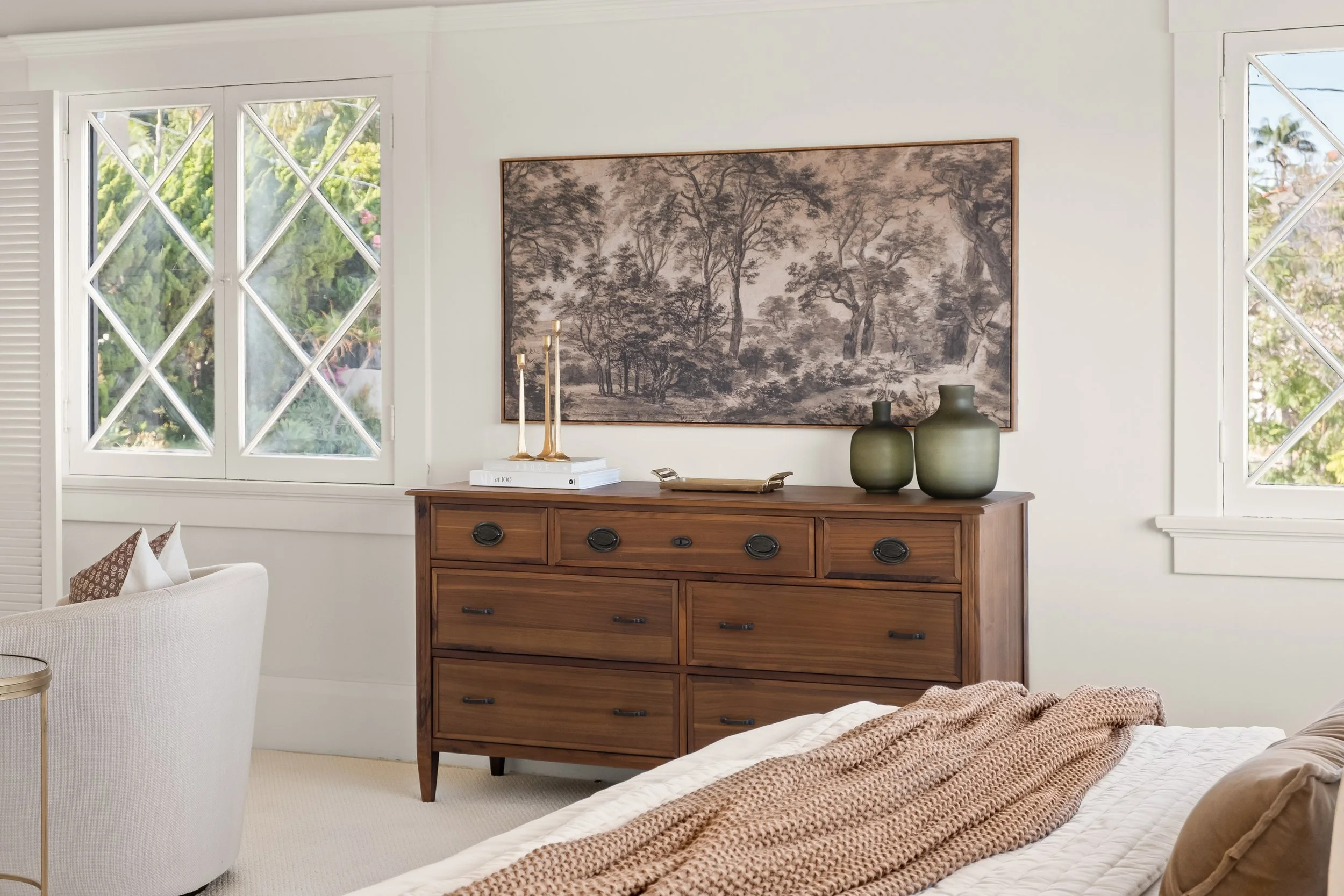 A cozy bedroom with white walls, large windows with a view of greenery, a wooden dresser with drawers, decorative vases, a stack of books, and wall art featuring trees.