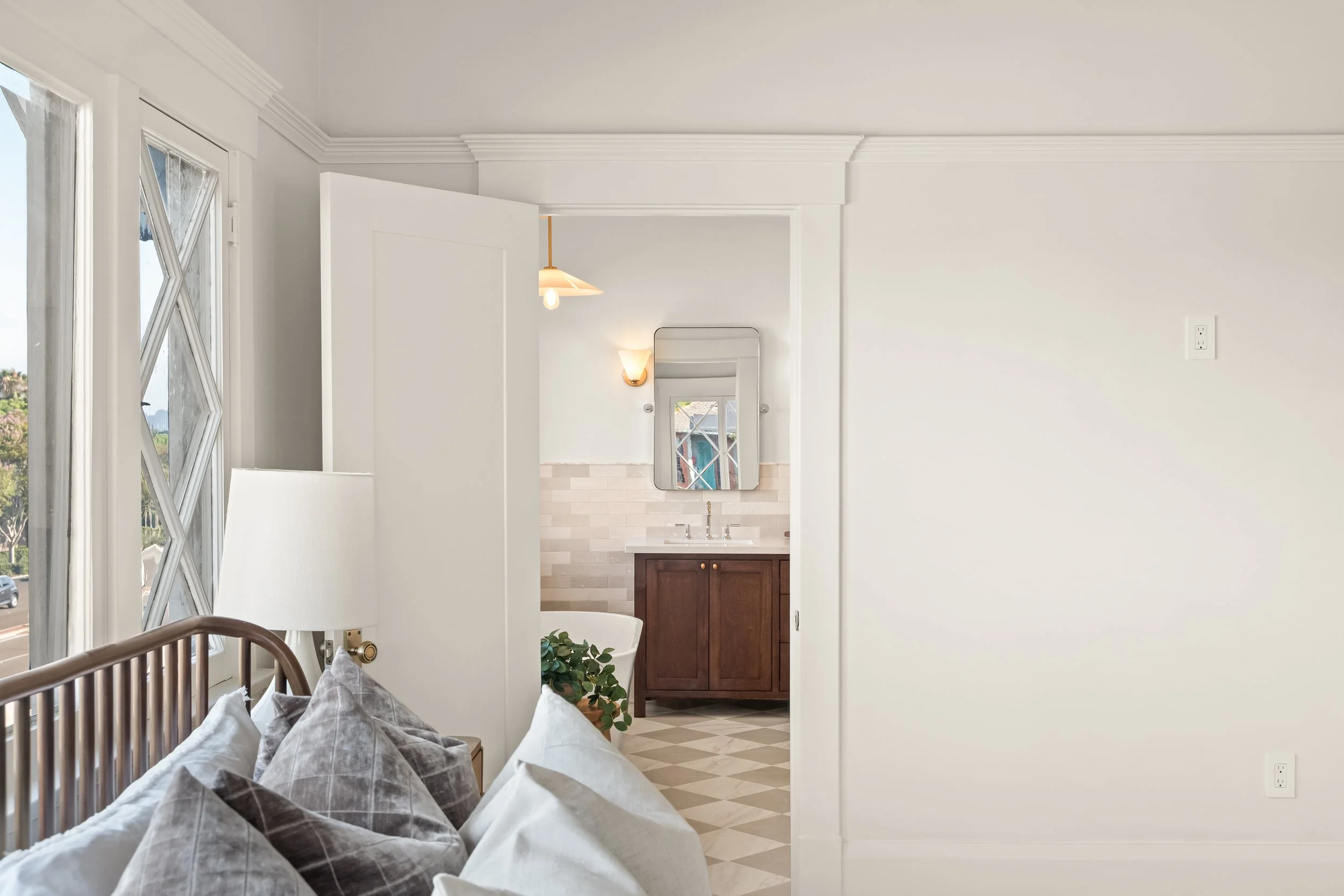 Interior of a bedroom with a view into a bathroom, featuring a bed with pillows, large windows, white walls, crown molding, a wooden vanity with a mirror, and a small plant on the floor.